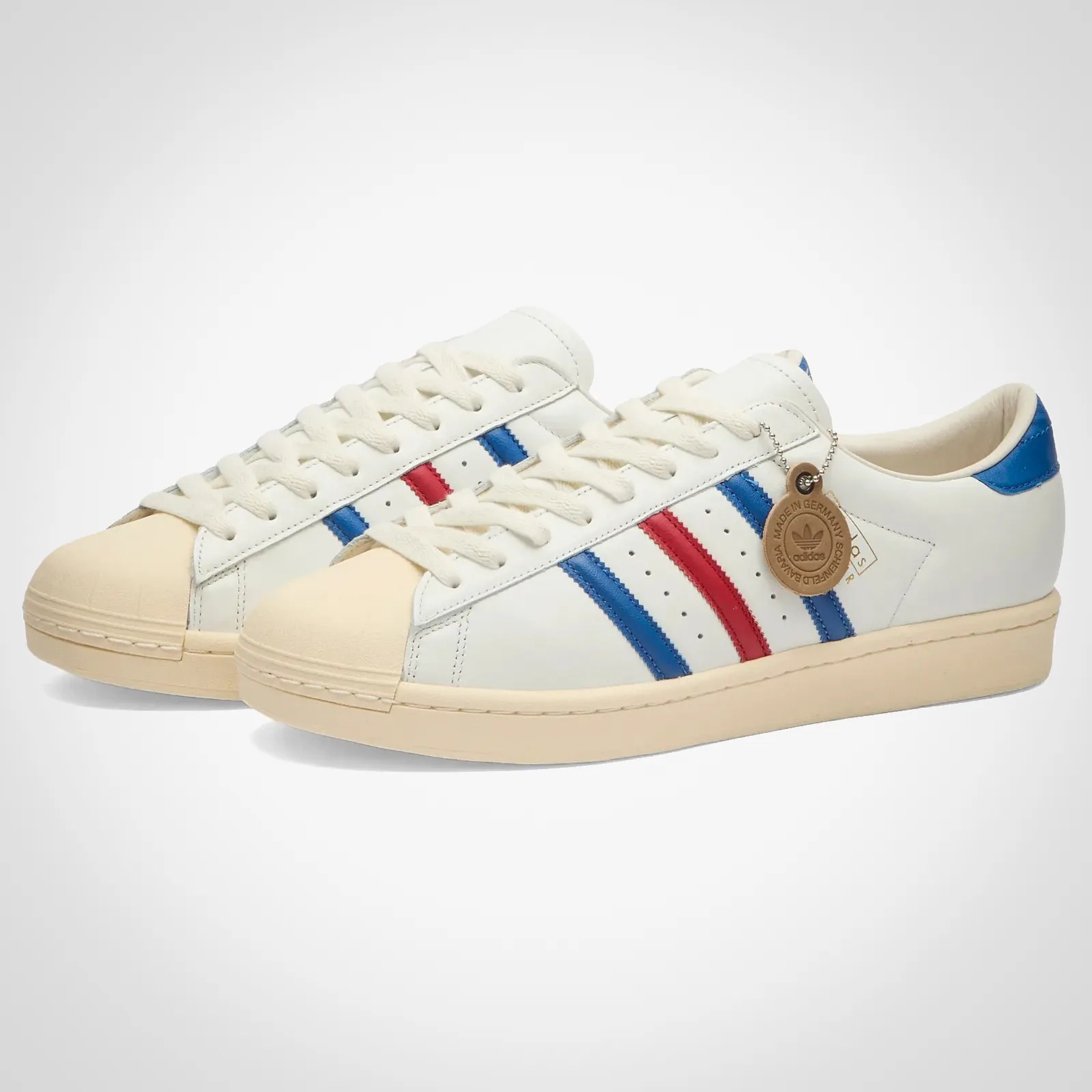 Pair of white Adidas sneakers with beige rubber toe caps and soles, featuring blue and red stripes on the sides. The shoes have white laces and a blue heel tab, with a round brown tag attached to one shoe.
