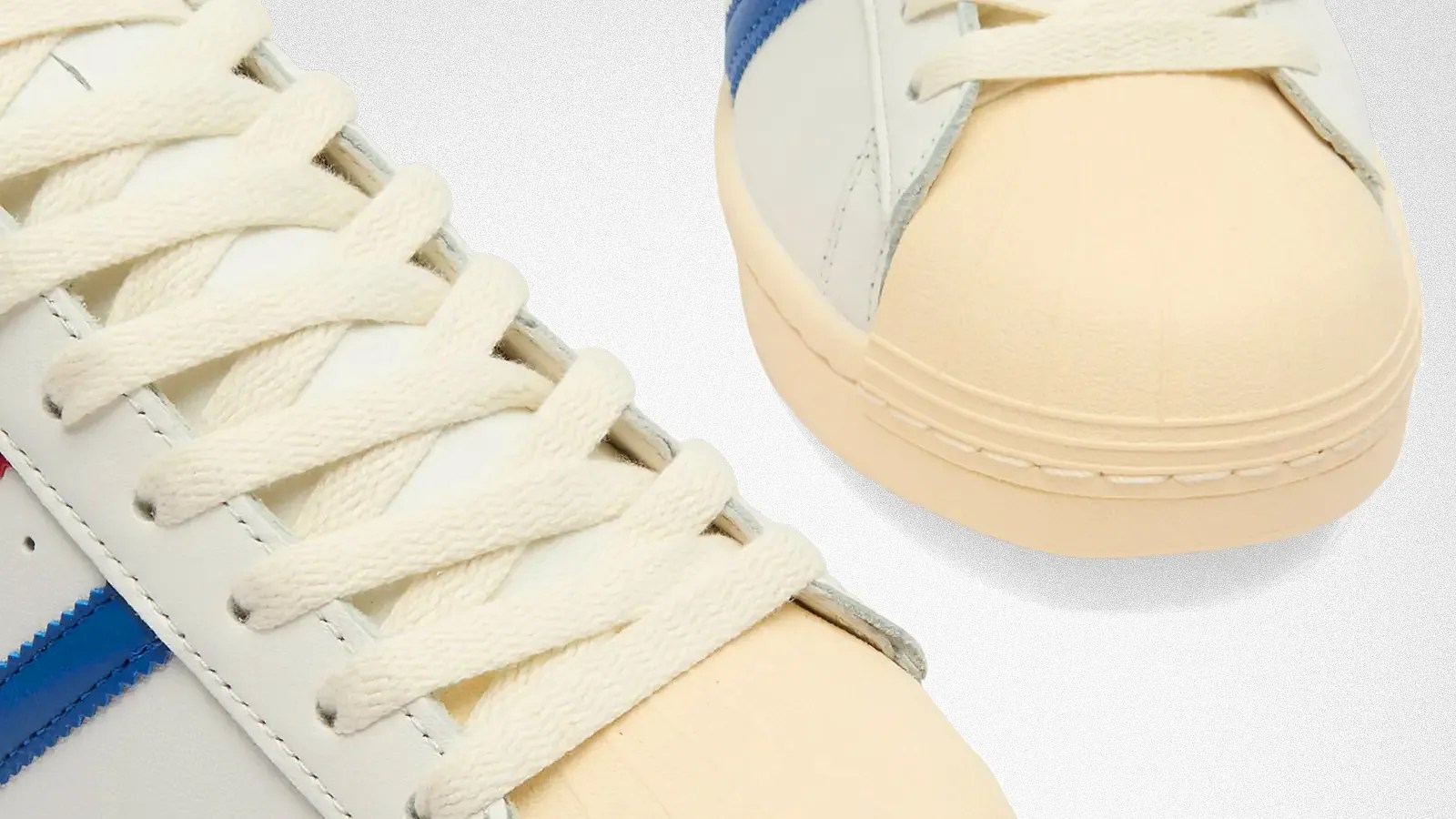 Close-up of the front part of a pair of sneakers featuring cream-colored laces, off-white rubber toe caps, and white leather uppers with blue and red accents. The stitching and texture of the materials are clearly visible.