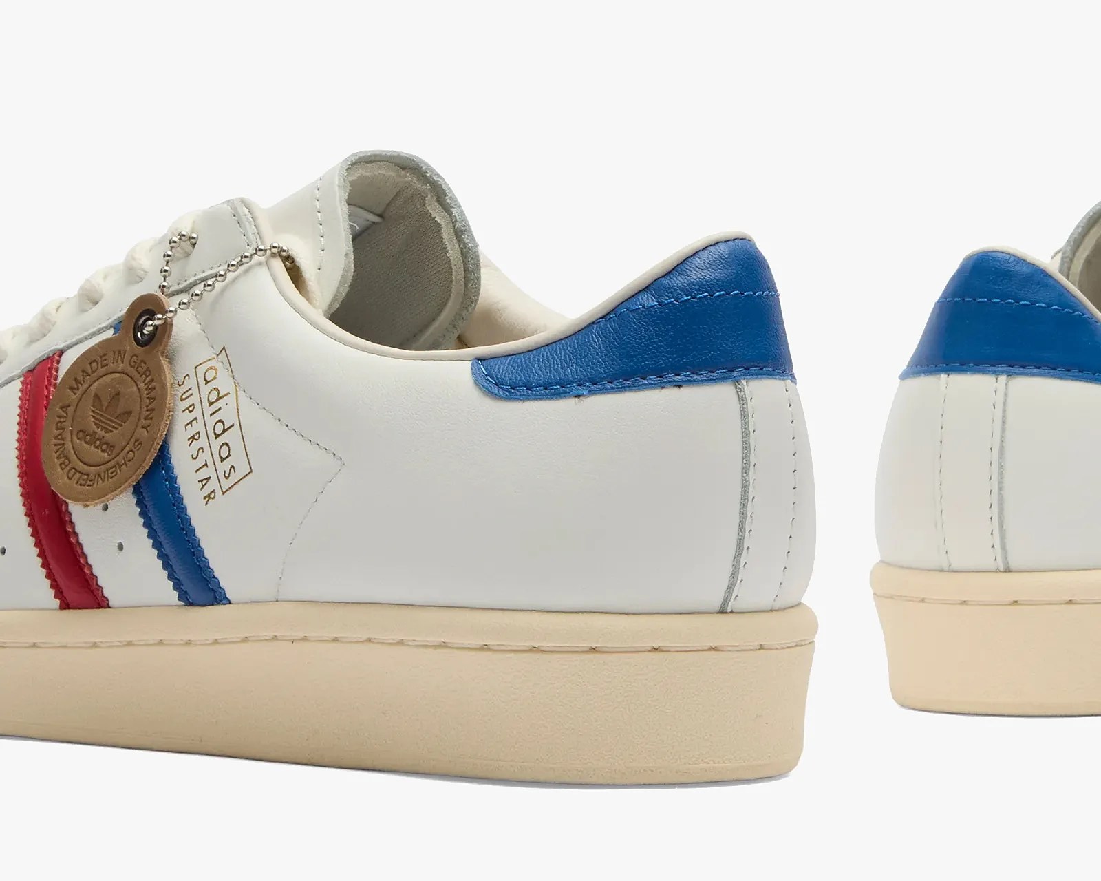 White Adidas Superstar sneakers with red and blue stripes on the side, blue heel tab, beige sole, and a brown leather tag attached near the laces that reads "Made in Germany." The Adidas logo and "Superstar" text are printed in gold on the side.
