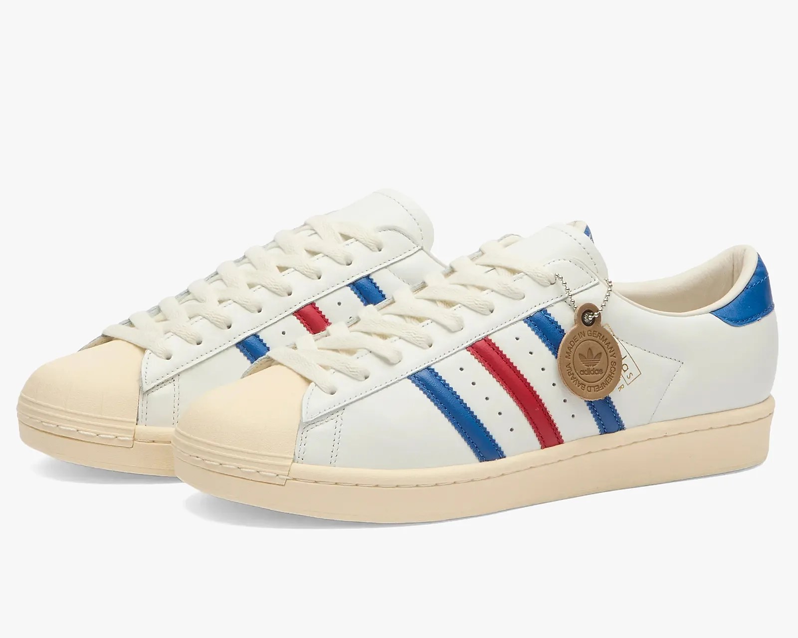 Pair of white Adidas sneakers with cream-colored rubber toe caps and soles, featuring blue and red stripes on the sides. The shoes have white laces and a blue heel tab, with a round brown tag attached to one shoe that reads "Made in Germany" and "Adidas.