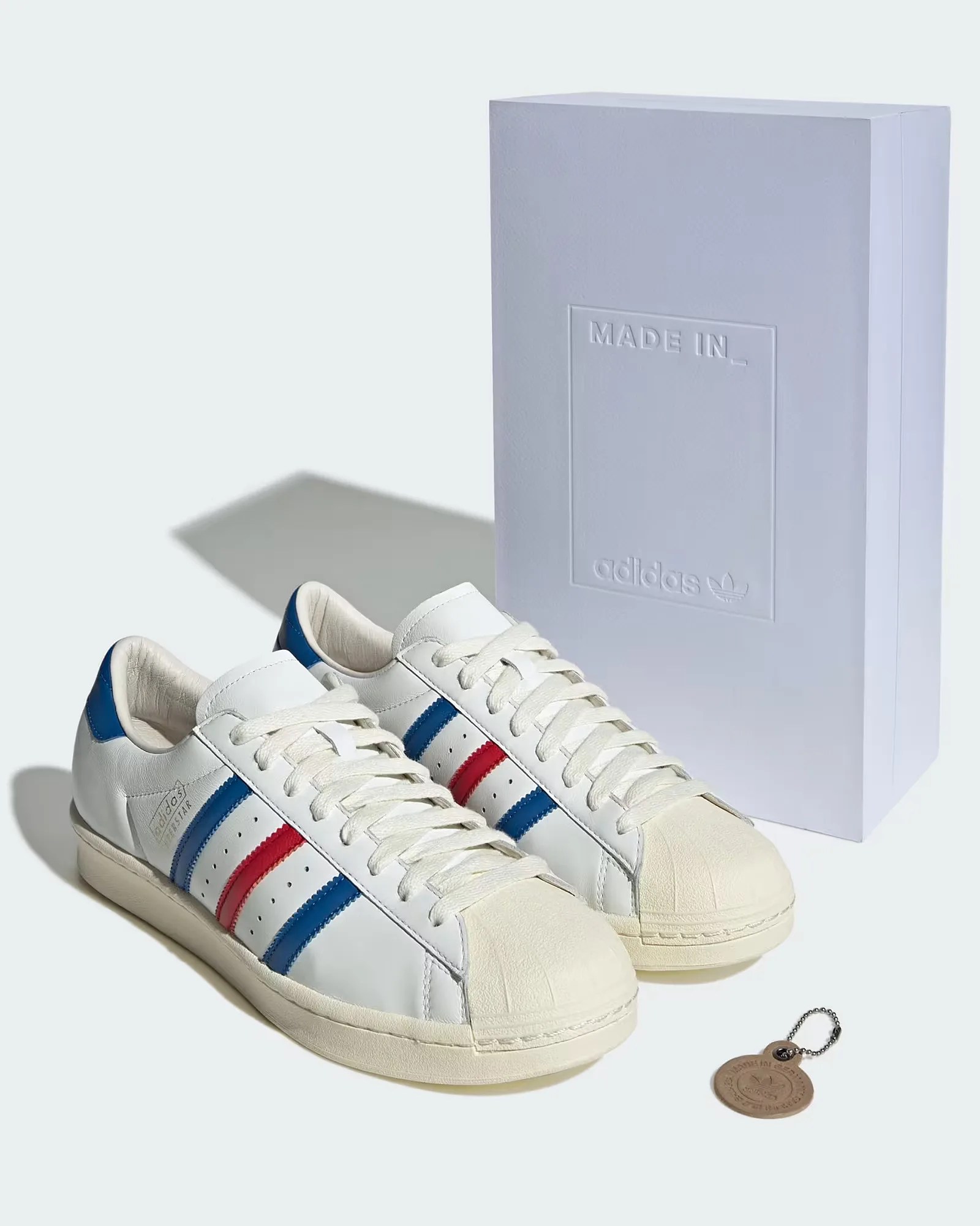 Pair of white Adidas sneakers with blue and red stripes, featuring a cream-colored shell toe and sole. The shoes are positioned in front of a tall white box embossed with "MADE IN_" and the Adidas logo. A small round leather keychain with a metal ball chain lies beside the shoes.