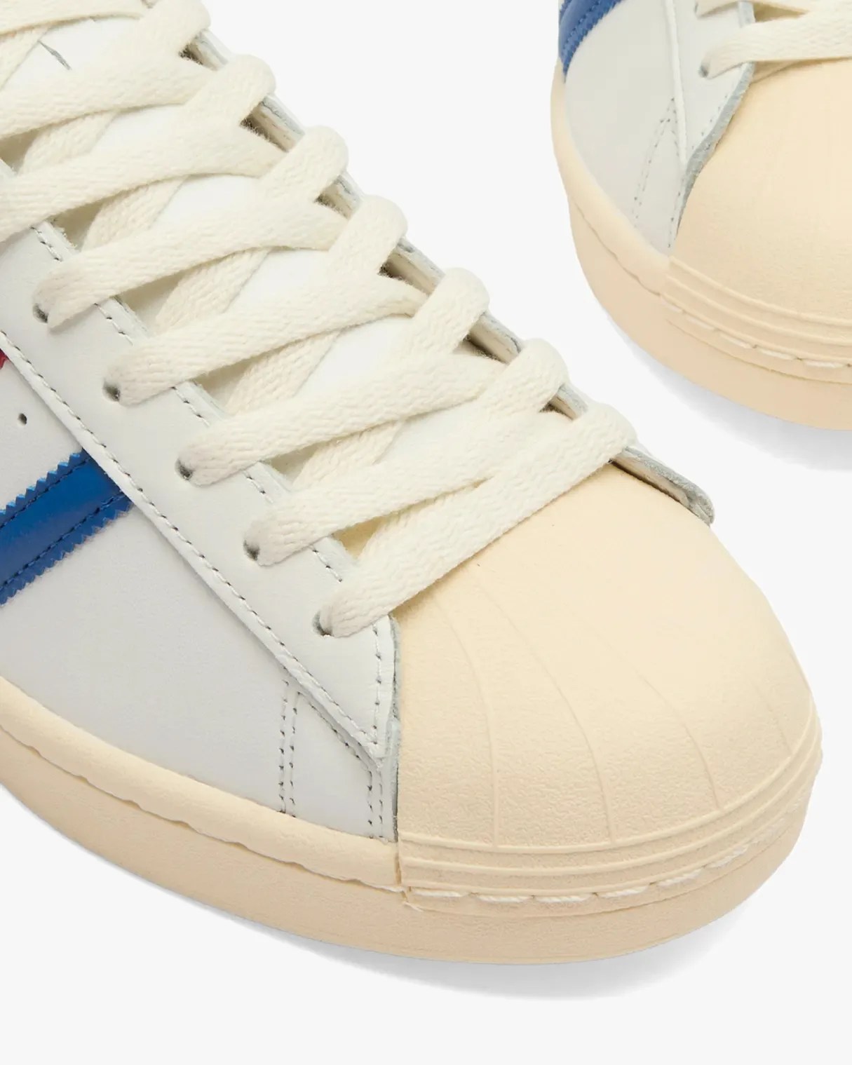 Close-up of a sneaker featuring off-white laces, a cream-colored rubber shell toe, and a white leather upper with a blue stripe detail. The sole is also cream-colored, matching the toe.