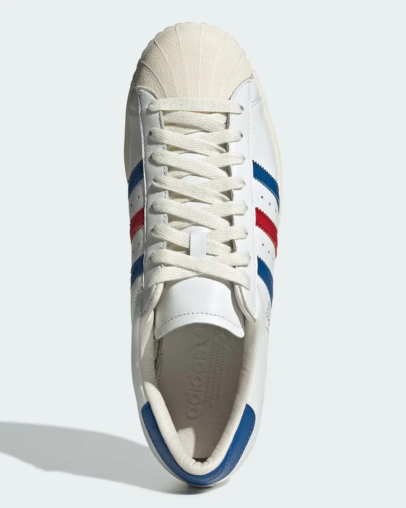 White Adidas sneaker with a shell toe design, featuring blue and red stripes on the sides. The shoe has white laces and a blue heel tab. The Adidas logo is embossed on the insole.