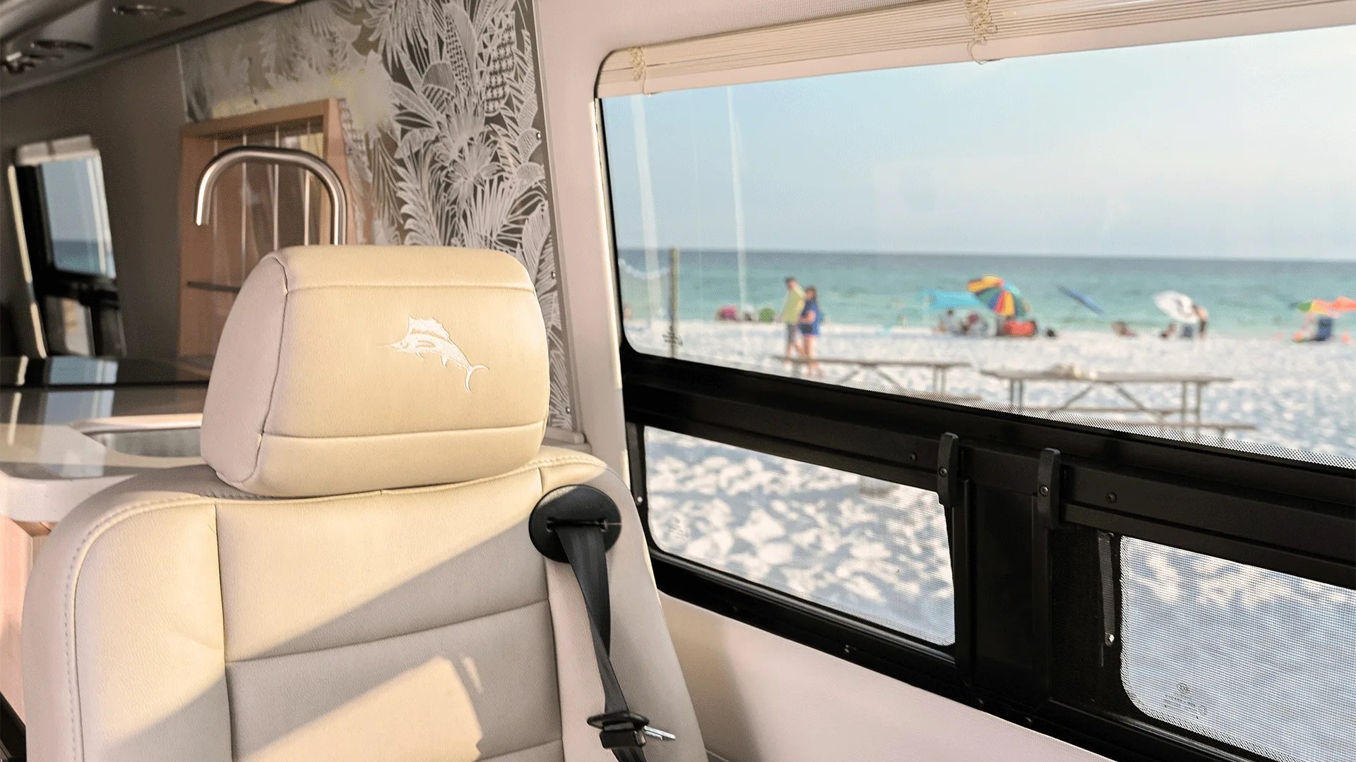 Beige leather seat with a marlin fish logo embroidered on the headrest inside a vehicle, positioned next to a window showing a beach scene with sand, ocean, and people under colorful umbrellas. The interior also features a kitchen sink and patterned wallpaper.