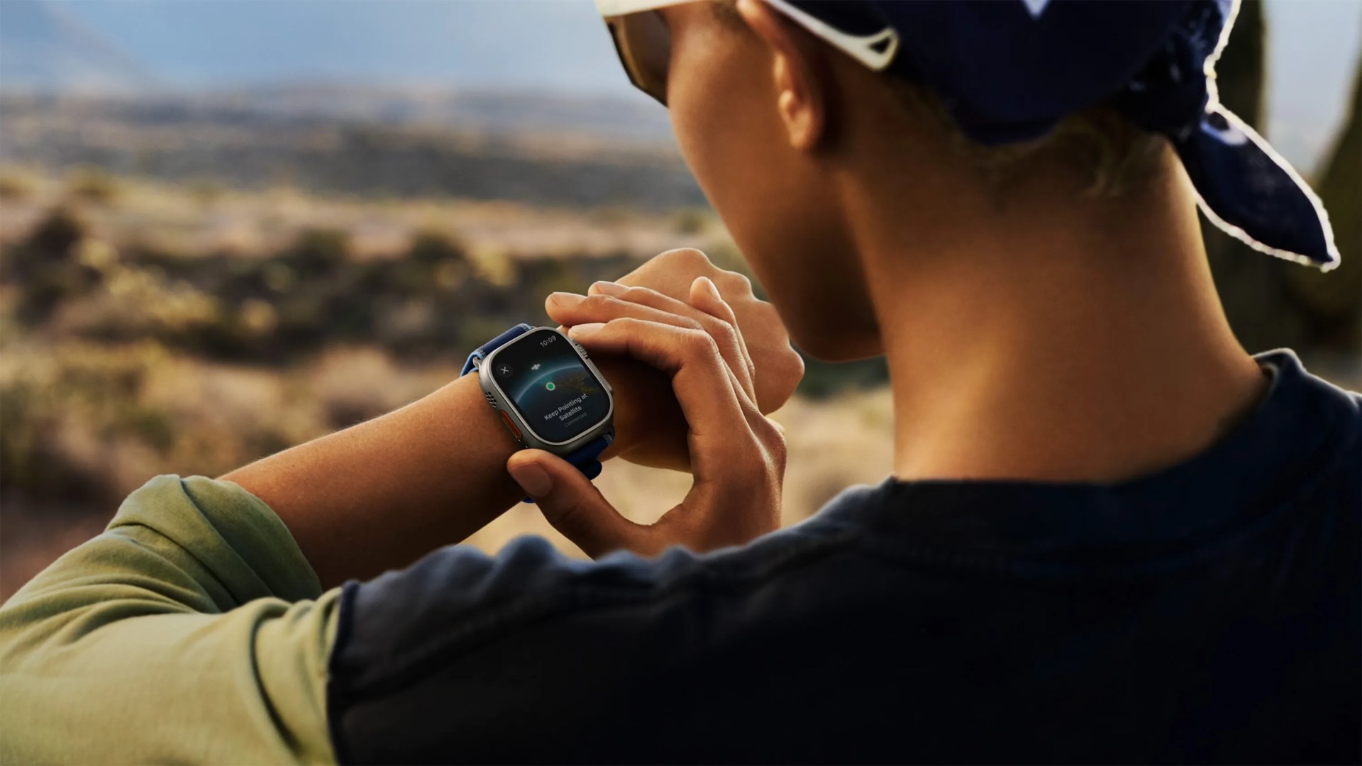 A person wearing a dark blue bandana and sunglasses is outdoors, looking at a smartwatch on their left wrist. The smartwatch screen displays a satellite connection interface with the text "Keep Pointing at Satellite." The person is using their right hand to interact with the watch. The background is a blurred natural landscape with muted colors.