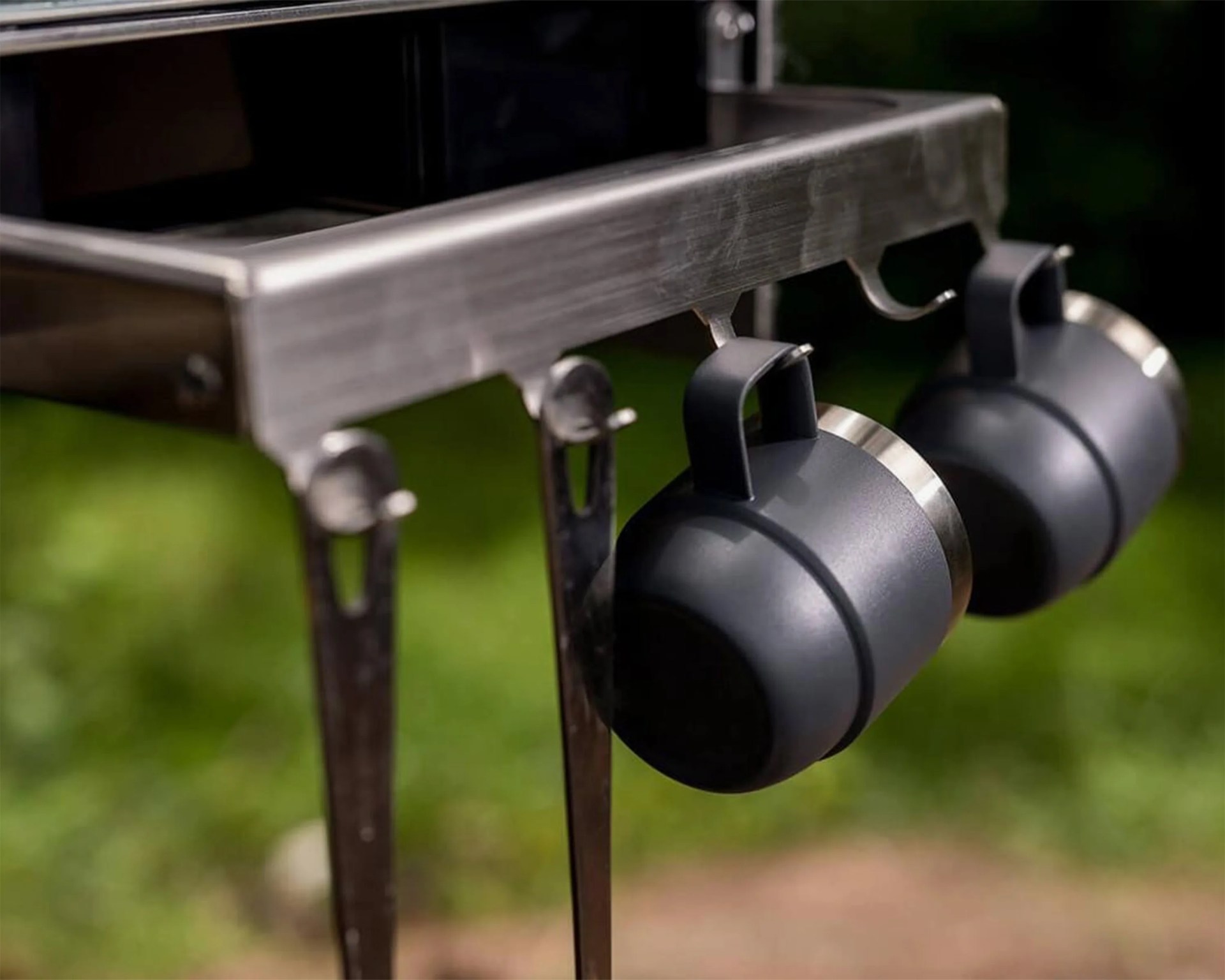 Two black metal mugs with handles hanging from hooks on a stainless steel rack, with a blurred green outdoor background.
