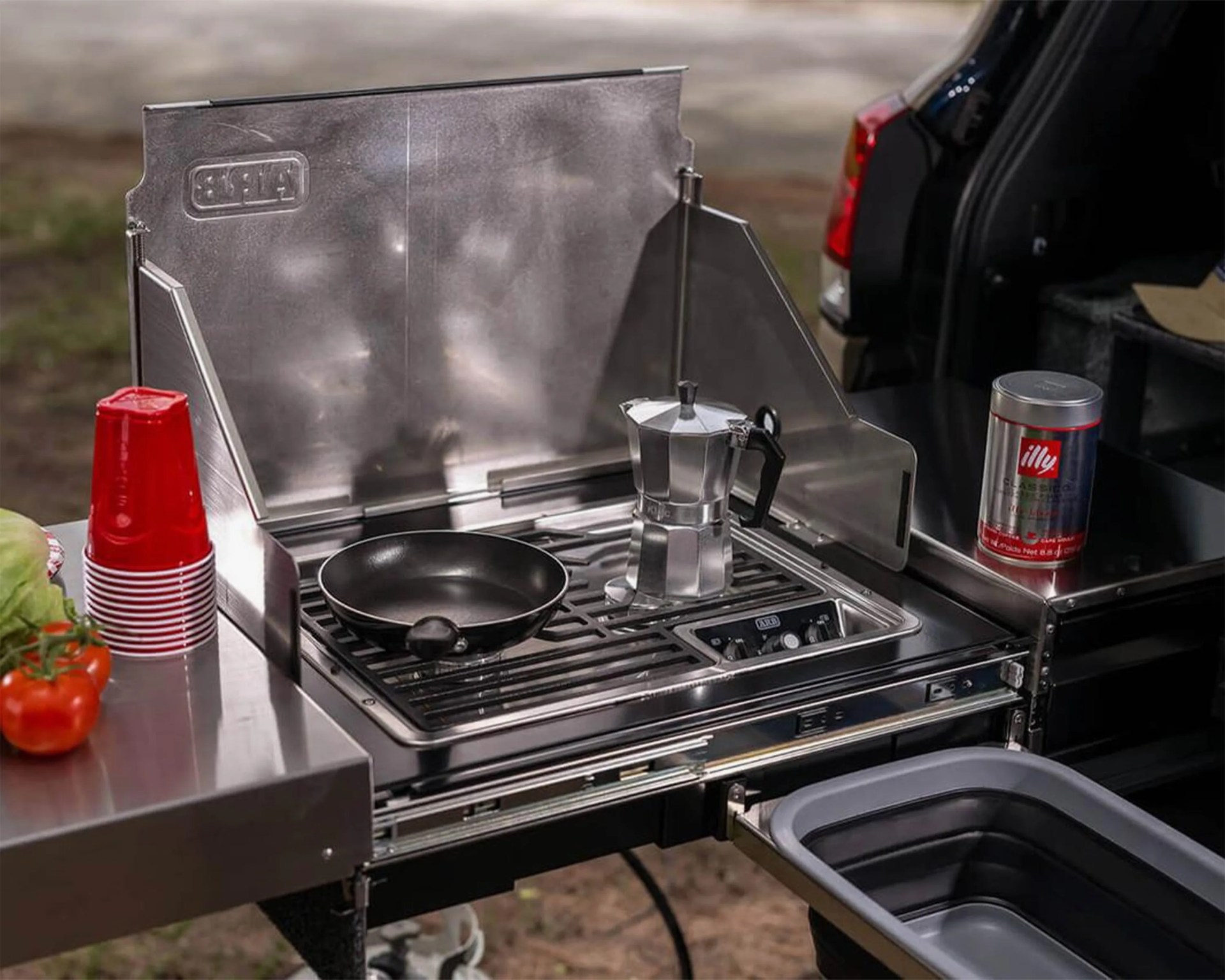 A portable stainless steel camping stove with two burners, one holding a black frying pan and the other a silver stovetop espresso maker. To the left, there is a stack of red plastic cups and some fresh vegetables including tomatoes and lettuce. To the right, a can of illy coffee sits on the stove's side surface, and a collapsible gray sink basin is pulled out below. The setup is attached to the back of a vehicle.