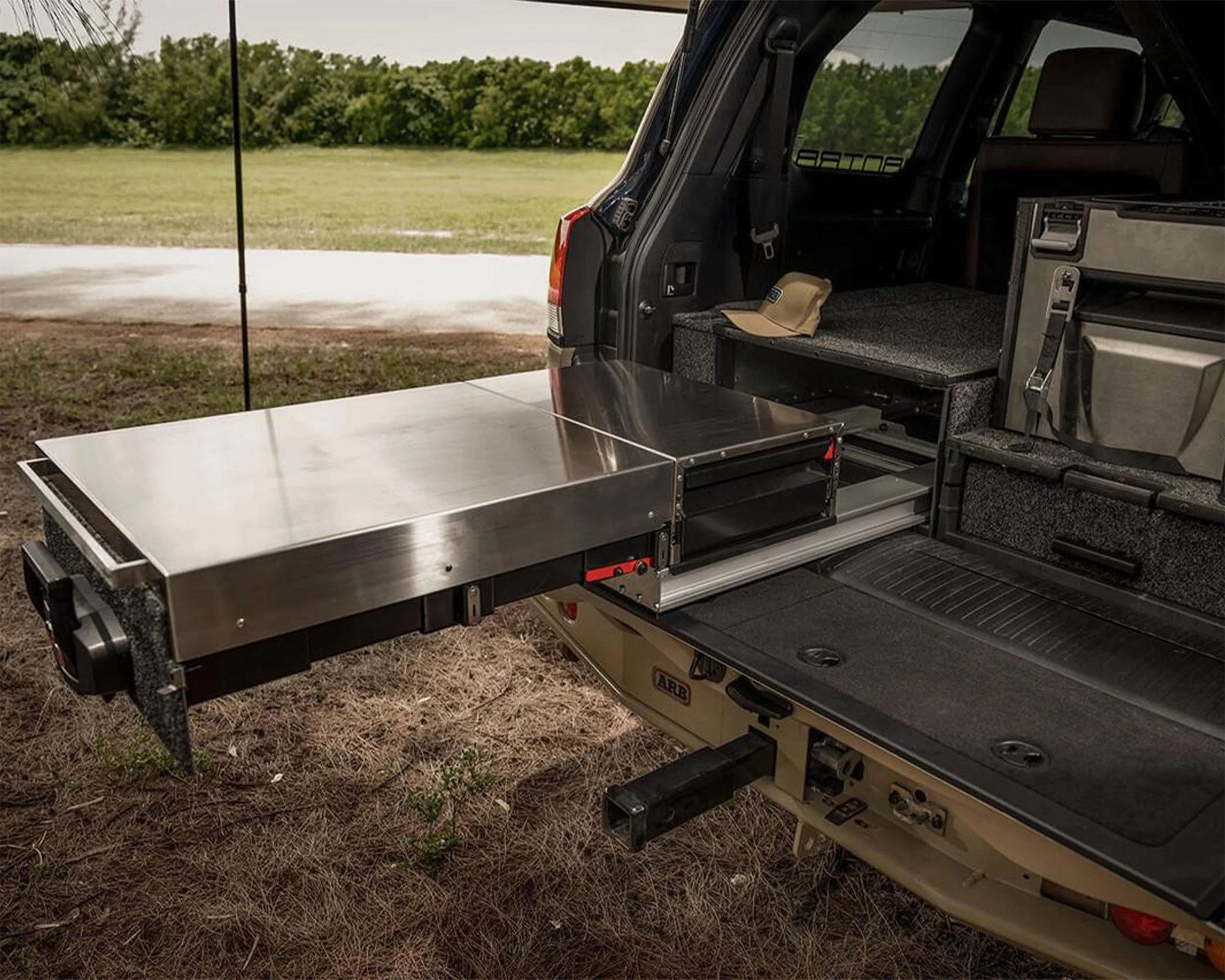 A metal sliding drawer system installed in the back of an SUV, partially extended. The drawer has a stainless steel top surface and is mounted on a carpeted platform inside the vehicle. A tan baseball cap rests on the platform next to the drawer. The vehicle's tailgate is open, revealing the interior setup.