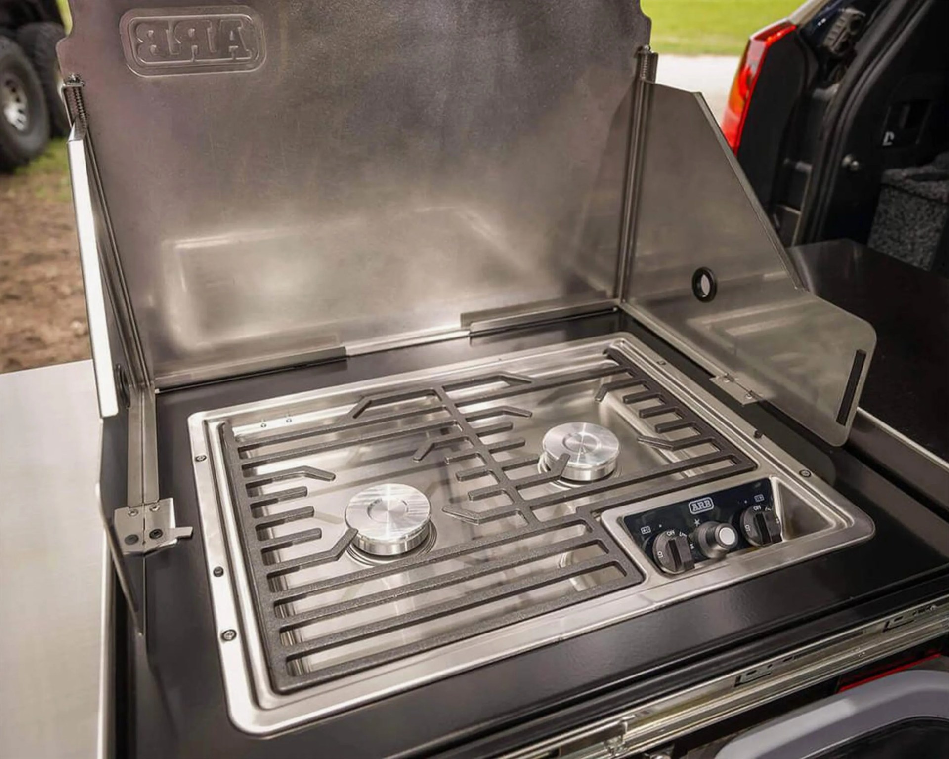 Stainless steel two-burner outdoor gas stove with control knobs on the right side, mounted on a black surface with a hinged metal wind guard around the back and sides. The stove has a sturdy metal grate over the burners and the brand "ARB" embossed on the inside of the wind guard.