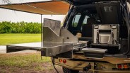 A rear view of an off-road vehicle with a slide-out stainless steel kitchen setup. The kitchen includes a flat work surface, a vertical panel, and a collapsible sink attached below the slide-out platform. The vehicle's tailgate is open, revealing the kitchen system installed inside the cargo area. An awning extends from the vehicle's side, providing shade. The vehicle is parked on grass with green bushes in the background.