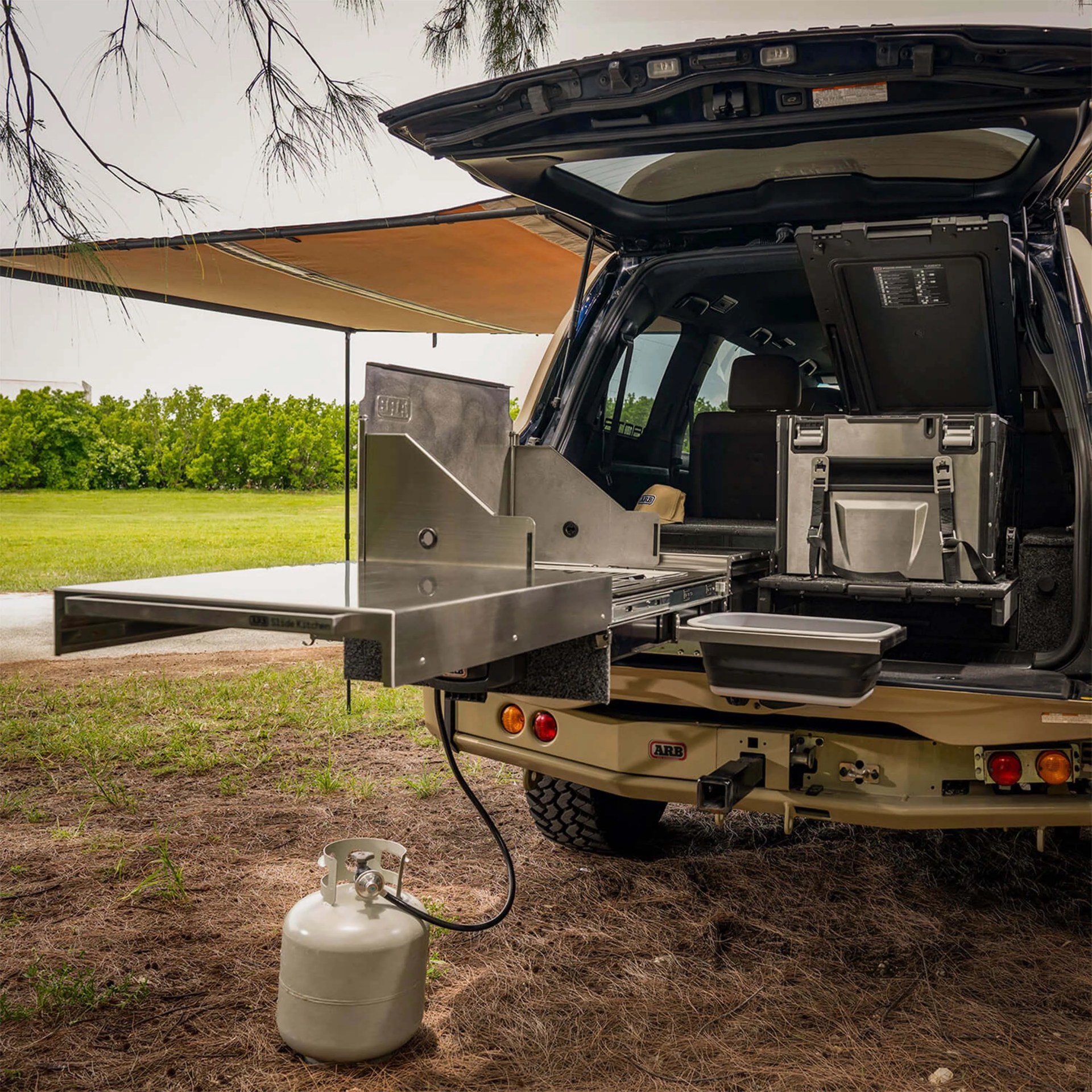 A beige off-road vehicle with its rear hatch open, revealing a slide-out stainless steel kitchen setup. The kitchen includes a flat work surface, a fold-up panel, and a connected propane tank on the ground. A retractable awning extends from the side of the vehicle, providing shade. The interior of the vehicle shows a storage box and seating. The scene is outdoors on a grassy area with trees in the background.