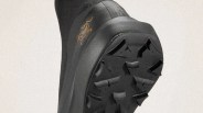 Close-up of the sole and heel area of a black shoe with a textured, rugged tread pattern. The shoe features a small, stylized orange logo on the side near the heel. The upper material appears to be a tightly woven fabric.