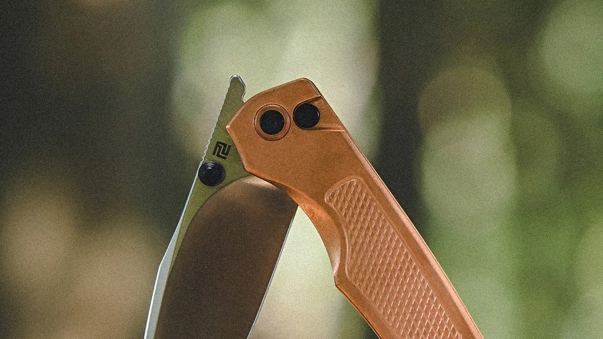 Close-up of a folding knife with a partially serrated stainless steel blade and a textured, copper-colored handle. The knife is shown in a partially open position against a blurred green and brown background.