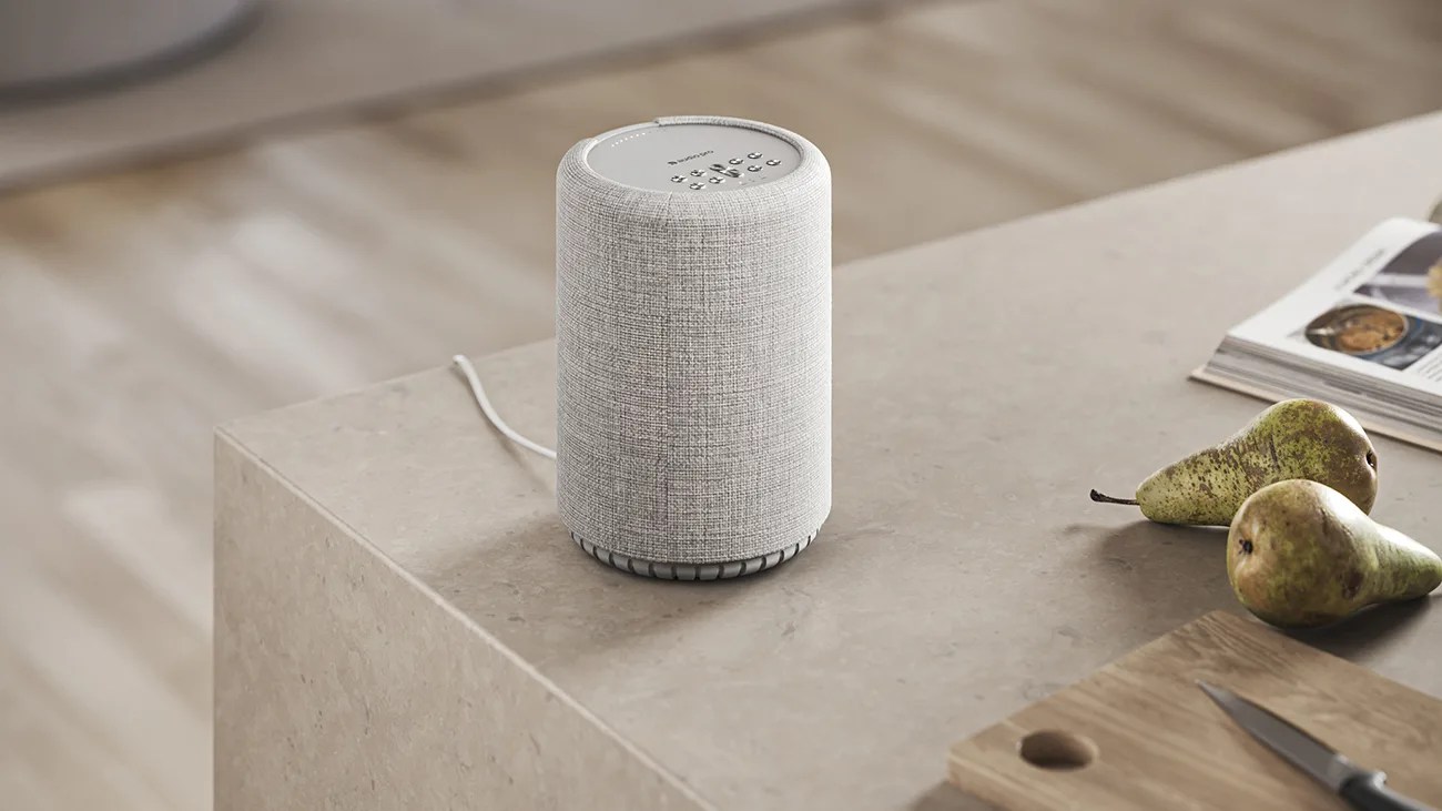 Gray fabric-covered smart speaker with control buttons on top, placed on a beige countertop near pears and a magazine.