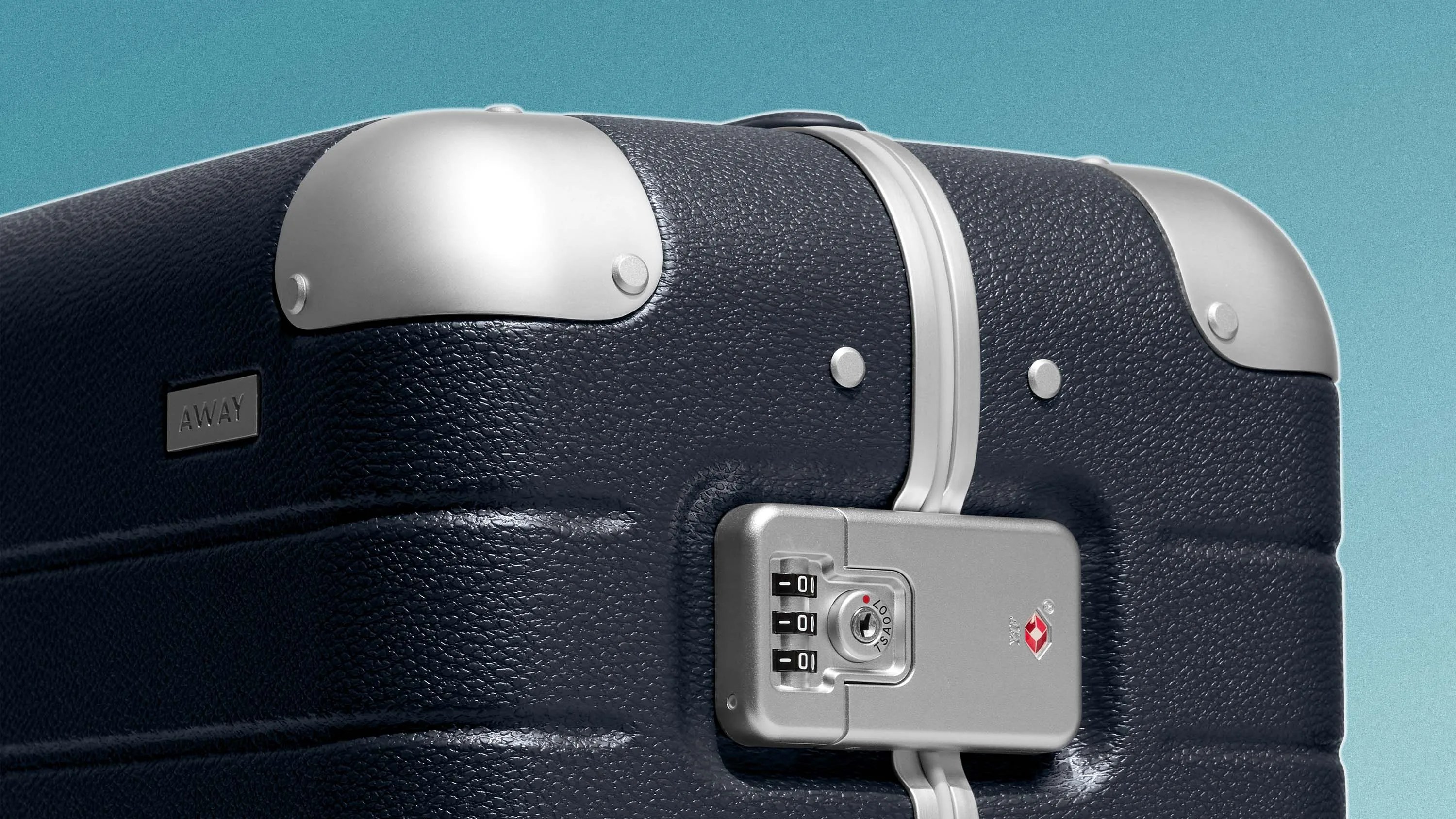 Close-up of a navy blue textured suitcase corner with silver metal corner protectors and a built-in TSA combination lock. The suitcase has a small black label with the brand name "AWAY" embossed on it. The background is a solid light blue color.