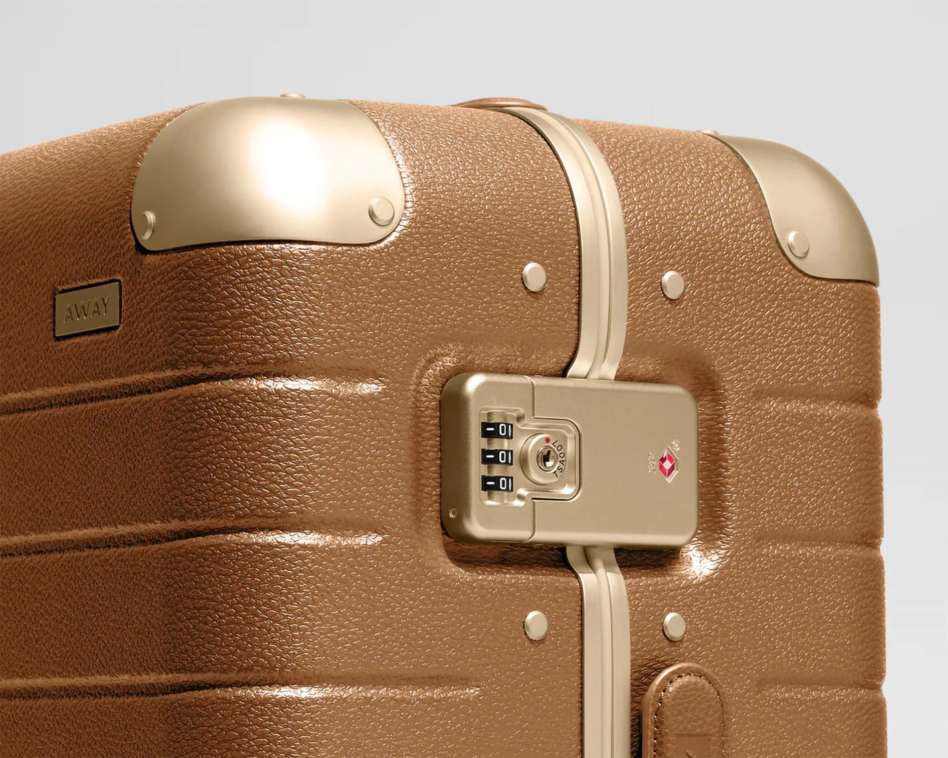 Close-up of a brown textured hard-shell suitcase corner with gold metal corner protectors and a built-in gold TSA combination lock. The suitcase has a small rectangular "AWAY" logo plate on the side.
