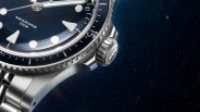 Close-up of a stainless steel wristwatch with a blue dial, featuring large triangular and circular hour markers, a rotating bezel with minute markings, and the text "AQUASCAPHE 200M" on the dial. The watch has a brushed metal bracelet and a prominent crown with a logo.