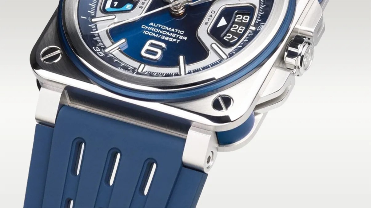 Close-up of a wristwatch with a blue dial and blue rubber strap. The watch features a square stainless steel case with visible screws at the corners, a crown on the right side, and a date display showing the 27th to 29th. The dial includes the text "AUTOMATIC CHRONOMETER 100M/325FT" and a prominent number 6. The strap has vertical cut-out details.