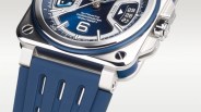 Close-up of a wristwatch with a blue dial and blue rubber strap. The watch features a square stainless steel case with visible screws at the corners, a crown on the right side, and a date display showing the 27th to 29th. The dial includes the text "AUTOMATIC CHRONOMETER 100M/325FT" and a prominent number 6. The strap has vertical cut-out details.