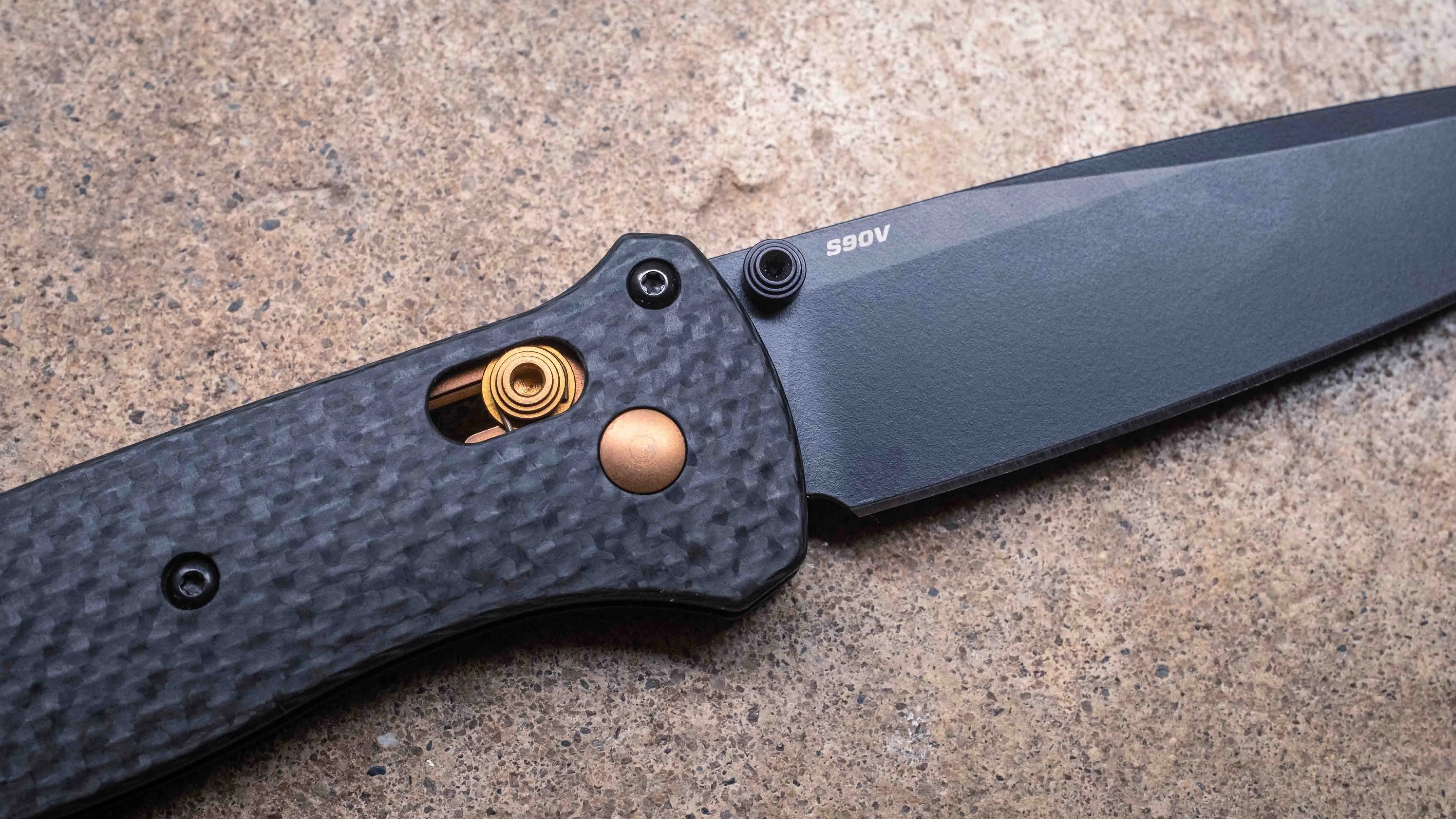 Black folding knife with a textured carbon fiber handle, a visible gold-colored spring mechanism, and a matte black blade marked "S90V." The knife is placed on a speckled stone surface.