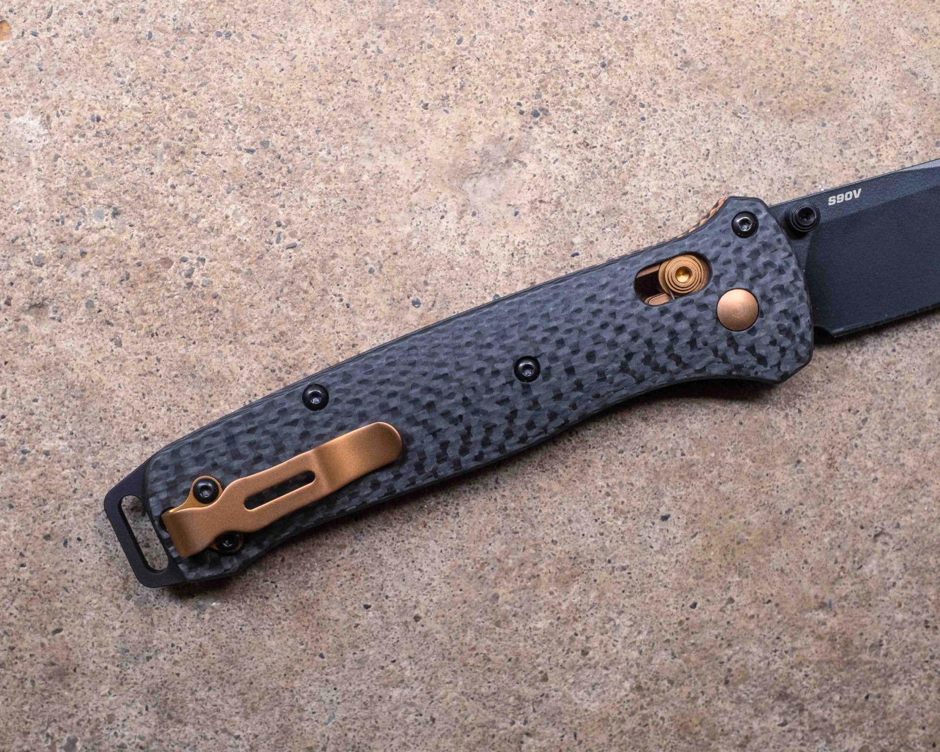 Black folding knife handle with a textured carbon fiber pattern, featuring a bronze-colored pocket clip, bronze pivot screw, and a visible bronze spring mechanism near the blade. The blade is partially visible and marked with "S90V." The knife is placed on a rough, beige stone surface.