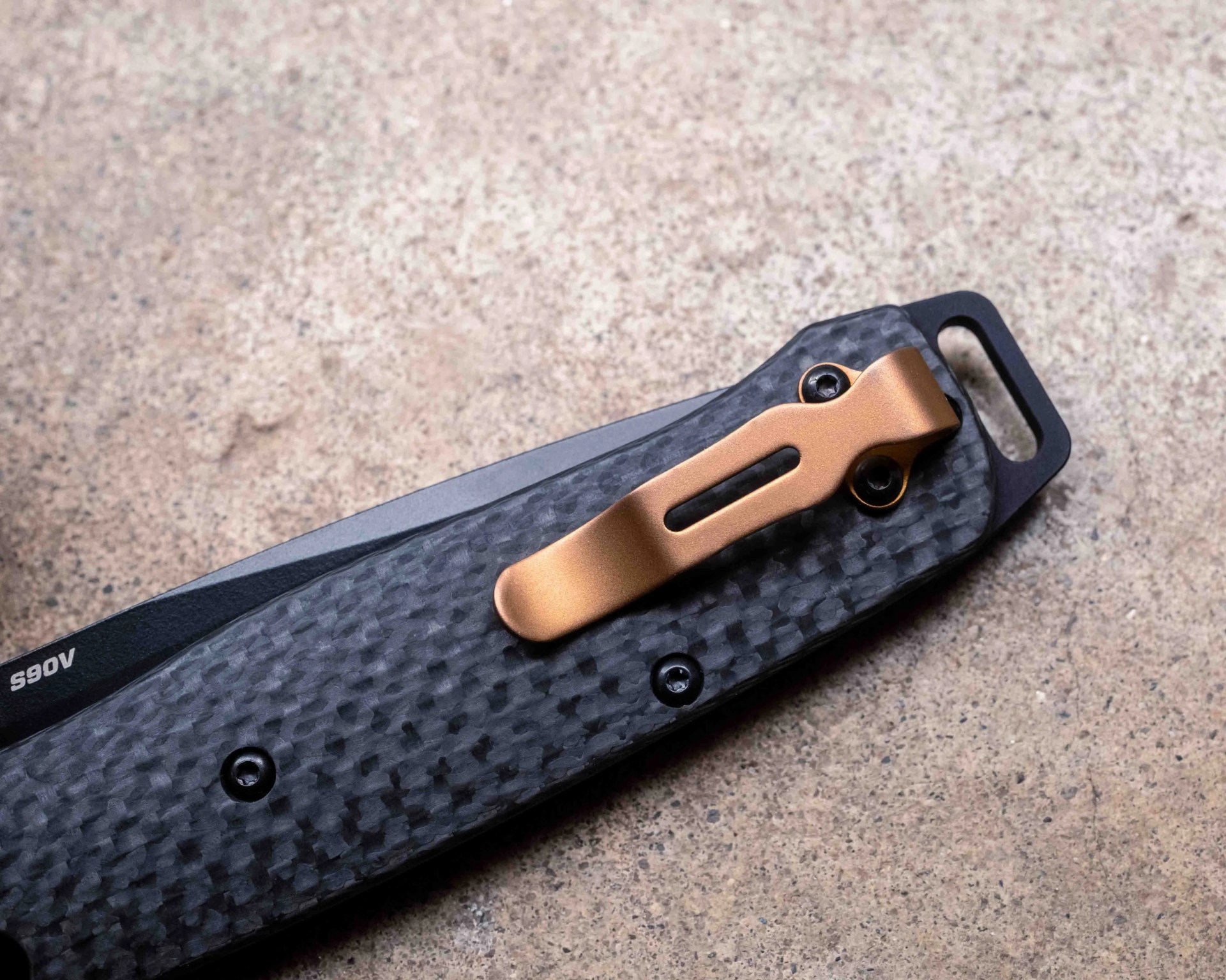 Close-up of a folding knife handle with a textured black carbon fiber grip, a bronze-colored metal pocket clip attached with two screws, and part of a black blade marked "S90V" visible. The background is a rough, light brown surface.