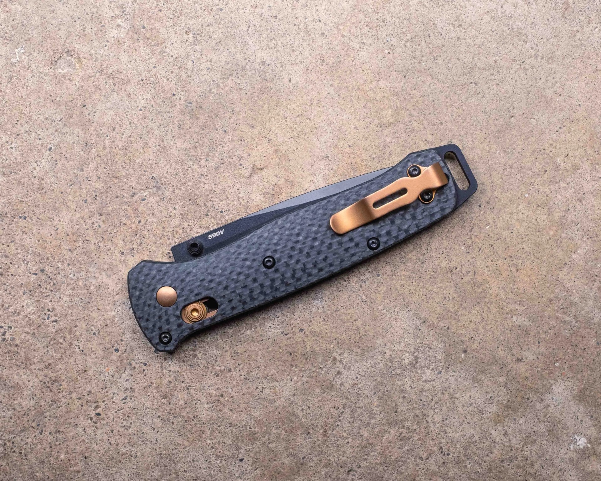 Folded pocket knife with a textured black handle, featuring a copper-colored clip and accents, resting on a rough beige surface. The blade is partially visible with the marking "A065.