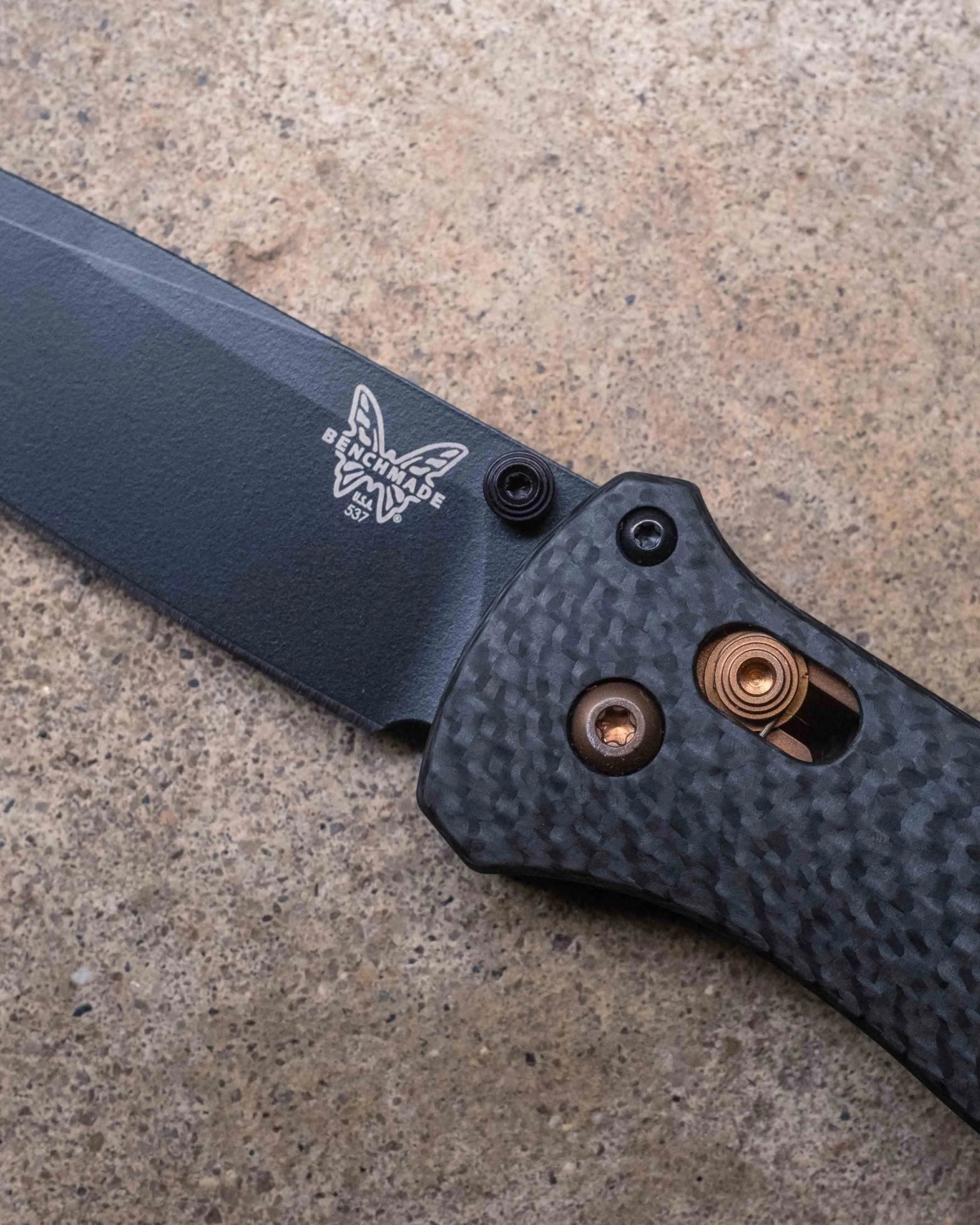 Close-up of a Benchmade folding knife showing the black blade with the Benchmade butterfly logo and model number 537. The handle has a textured, dark carbon fiber pattern with visible bronze-colored screws and a spring mechanism. The knife is placed on a speckled stone surface.