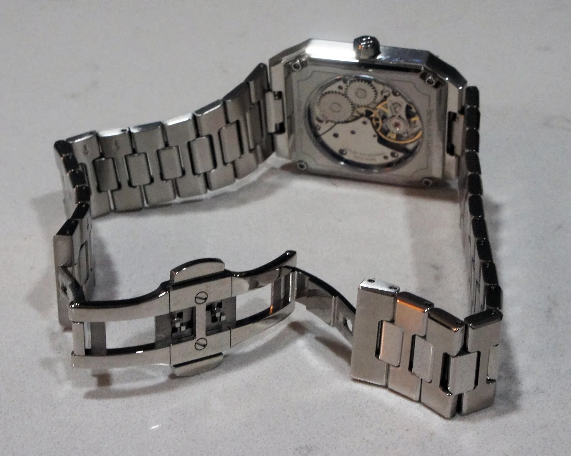 Silver metal wristwatch with a rectangular case and visible mechanical gears on the back, featuring a linked metal bracelet and an open clasp.