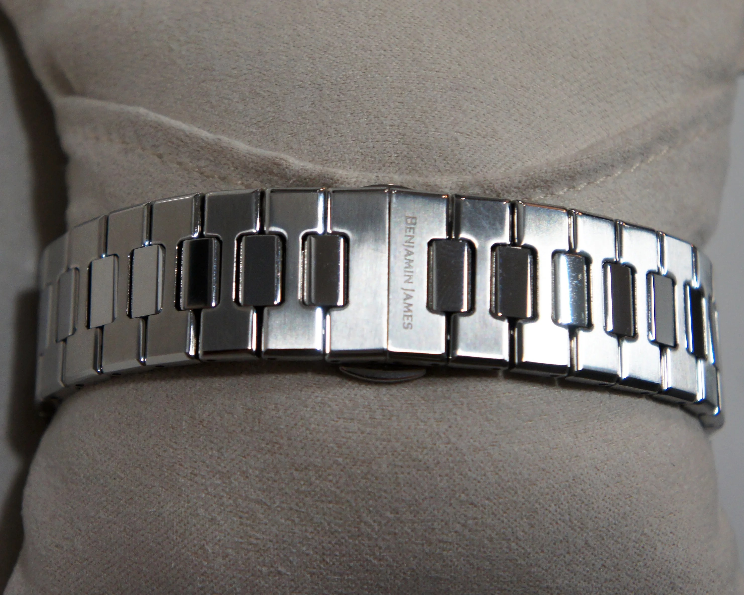 Silver metal watch band with rectangular links, featuring the engraved text "BENJAMIN JAMES" on the clasp, displayed on a beige fabric cushion.