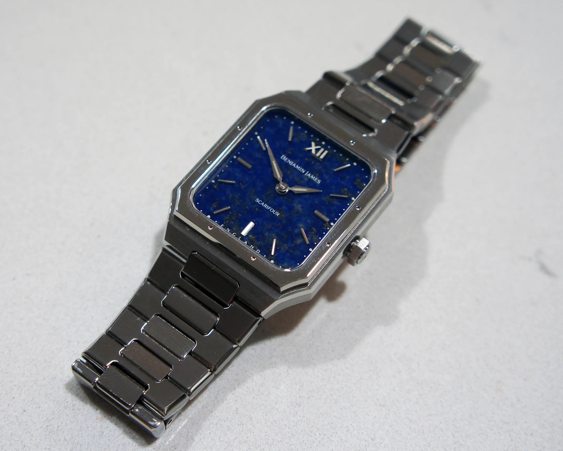 Square-faced wristwatch with a metallic silver band and a deep blue dial. The dial features silver hour markers, with a Roman numeral XII at the top, and the brand name "BENJAMIN JAMES" along with the model name "SCARIFOUR" printed on it. The watch has a polished silver bezel and a crown on the right side.