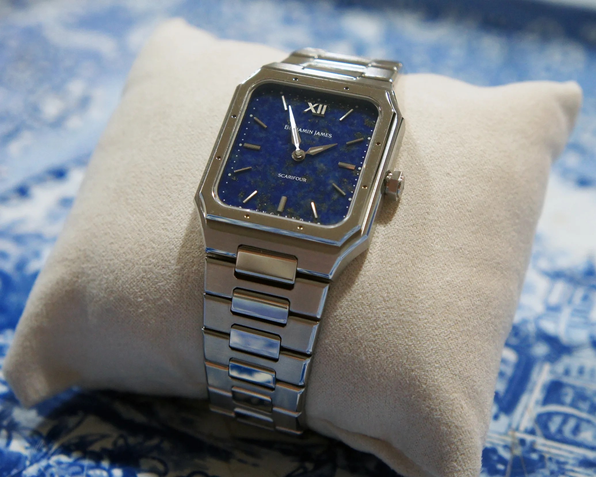 Square-faced wristwatch with a silver metal band and a deep blue dial featuring gold hour markers and hands, resting on a beige cushion. The watch face displays the brand name "Benjamin James" and the model name "Scarifour.
