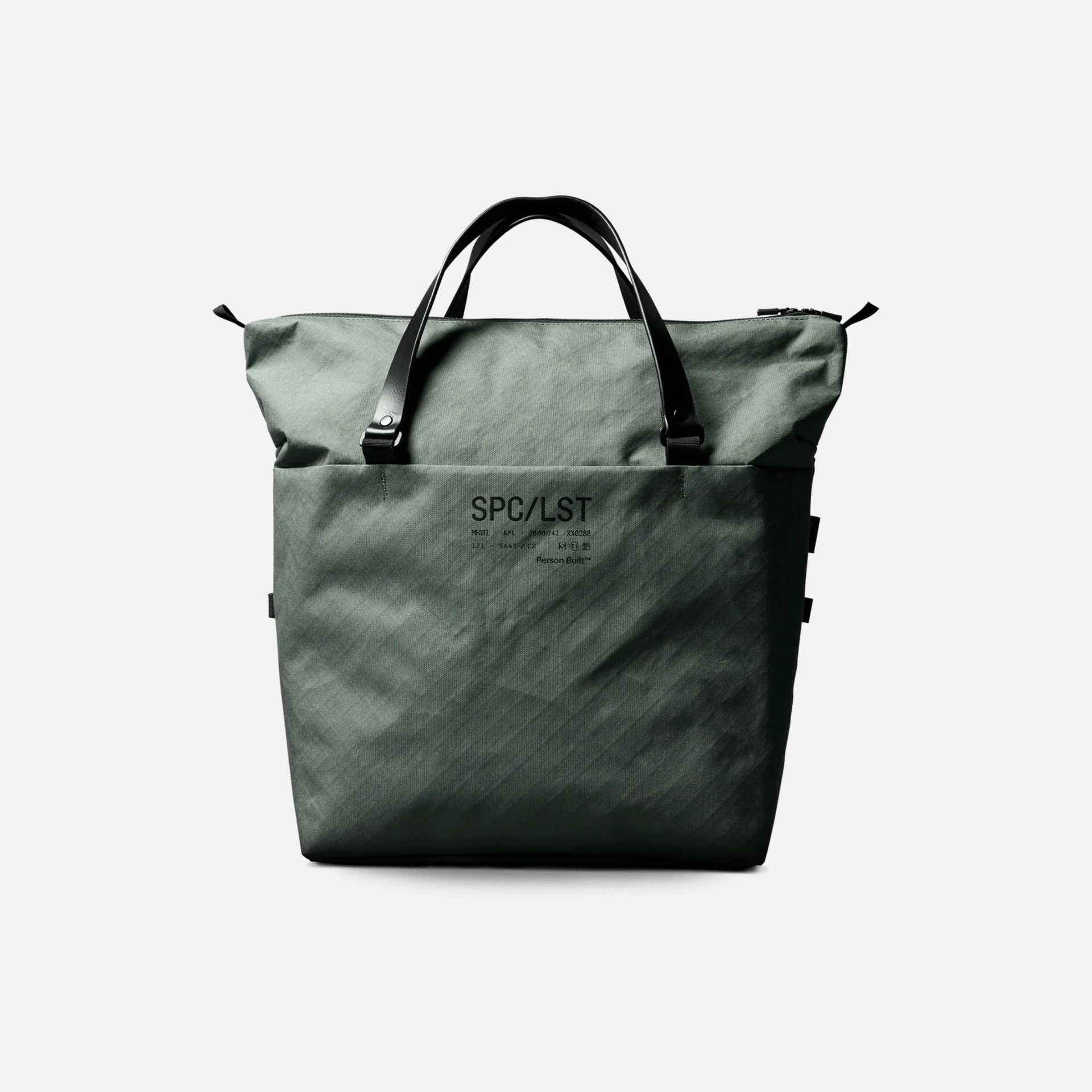 Green tote bag with black handles, featuring the text "SPC/LST" and additional small printed details on the front pocket. The bag has a minimalist design and a zipper closure at the top.
