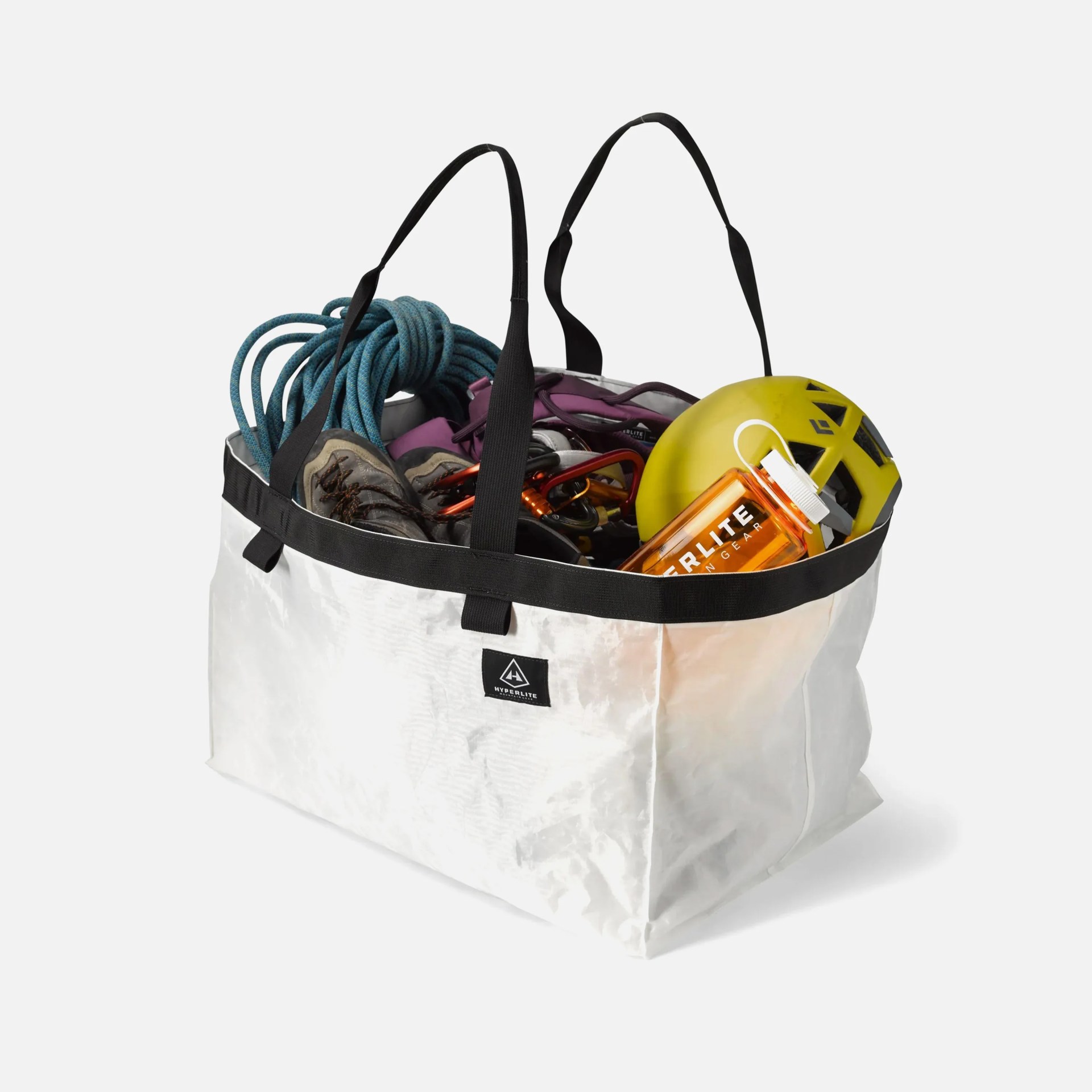 White rectangular gear bag with black handles and trim, filled with climbing equipment including a coiled blue rope, climbing shoes, carabiners, a yellow helmet, and an orange water bottle. The bag has a small black label with a white logo and text.