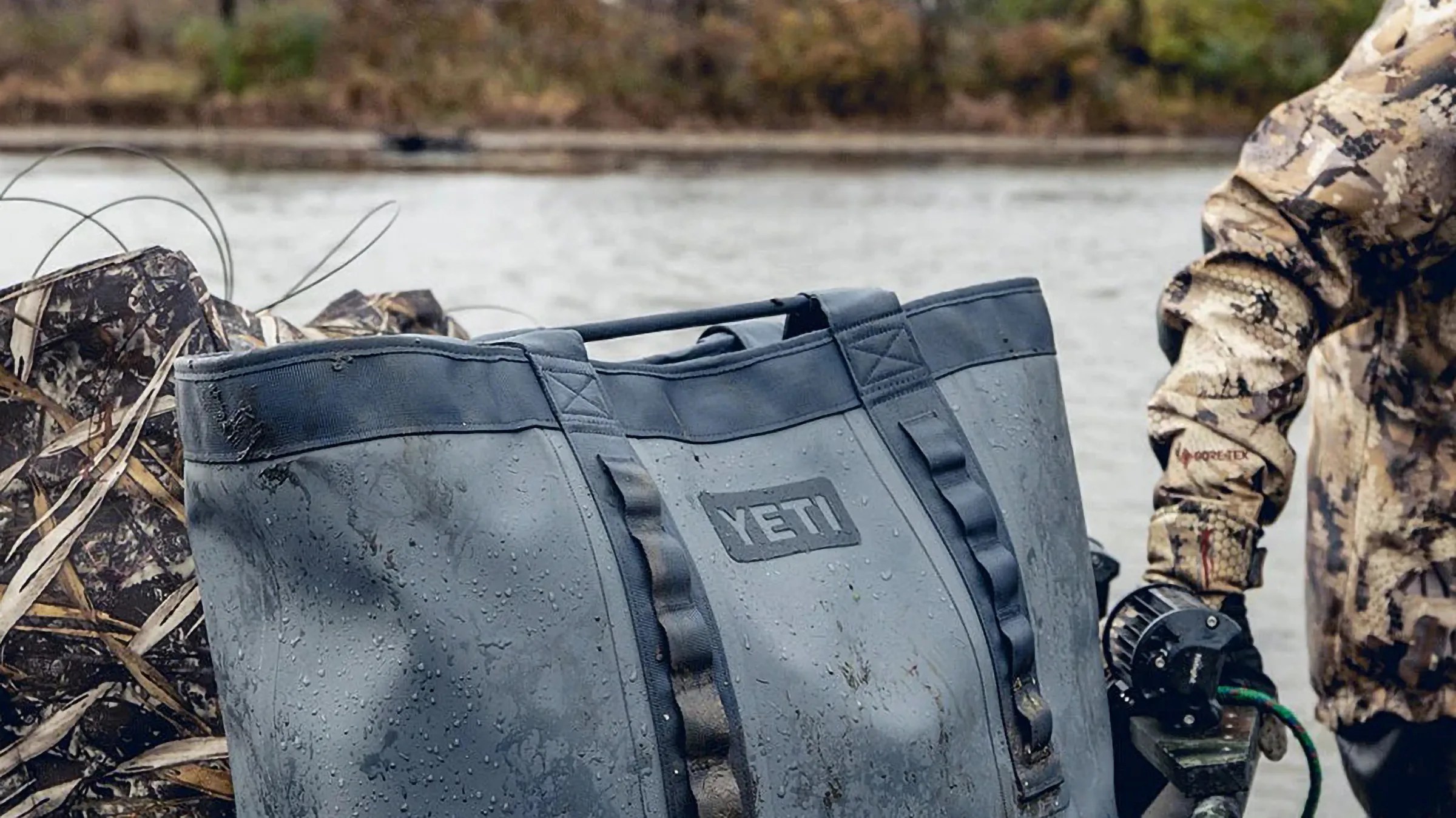 A large, gray YETI tote bag with water droplets on its surface, indicating it is wet. The bag has two sturdy handles and is positioned outdoors near a body of water. Part of a person wearing camouflage clothing and black gloves is visible on the right side of the image.