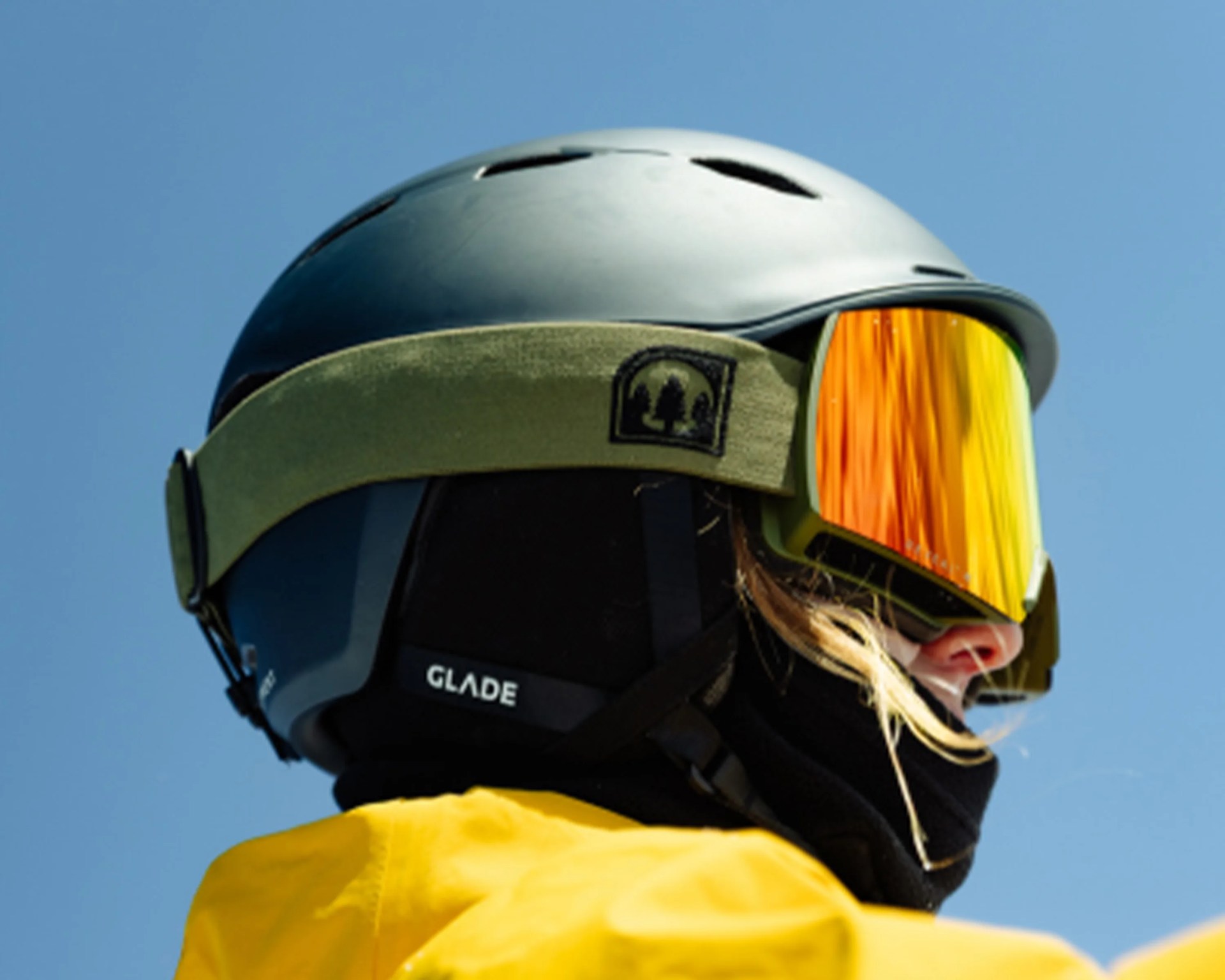 Side profile of a person wearing a matte black helmet with ventilation holes, reflective orange ski goggles with an olive green strap featuring a tree logo, a black face mask labeled "GLADE," and a bright yellow jacket against a clear blue sky.