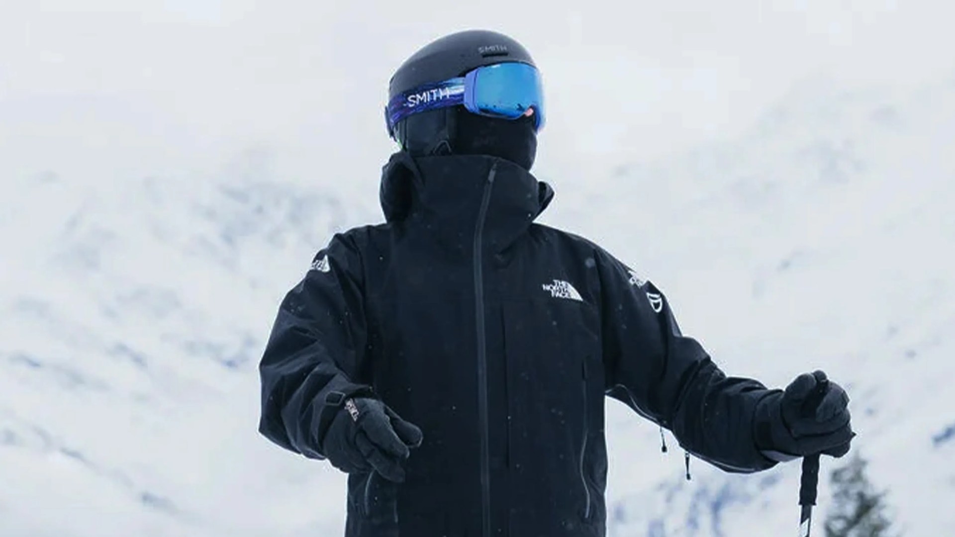 Person dressed in black winter gear, including a jacket, gloves, helmet, and blue ski goggles, standing in a snowy mountain environment. The jacket has "The North Face" logo on the chest and sleeve. The person is holding ski poles.