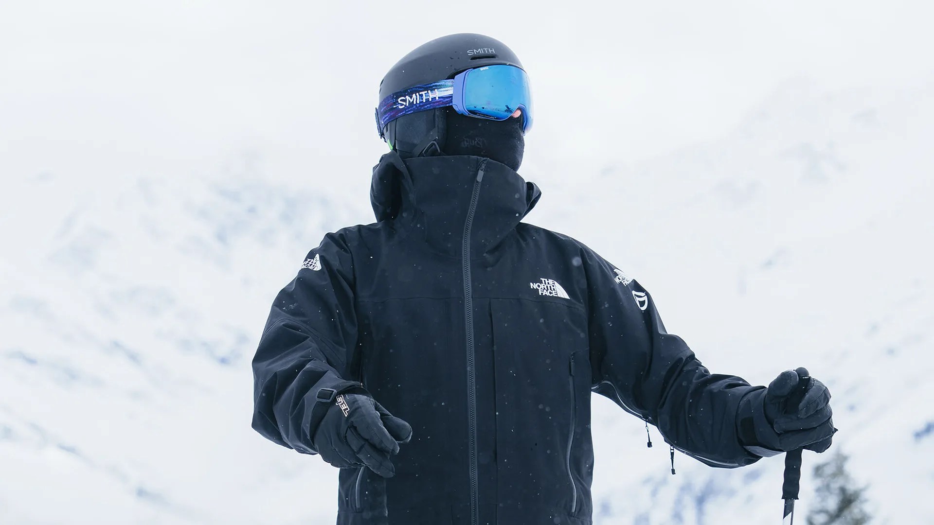 Person dressed in black winter gear, including a The North Face jacket, black gloves, a black helmet with "SMITH" written on it, and blue-tinted ski goggles, standing in a snowy mountain environment holding ski poles.