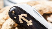 Close-up of a folding pocket knife with a polished wooden handle featuring a gold anchor emblem, and a partially visible stainless steel blade. The background includes coiled rope, suggesting a nautical theme.