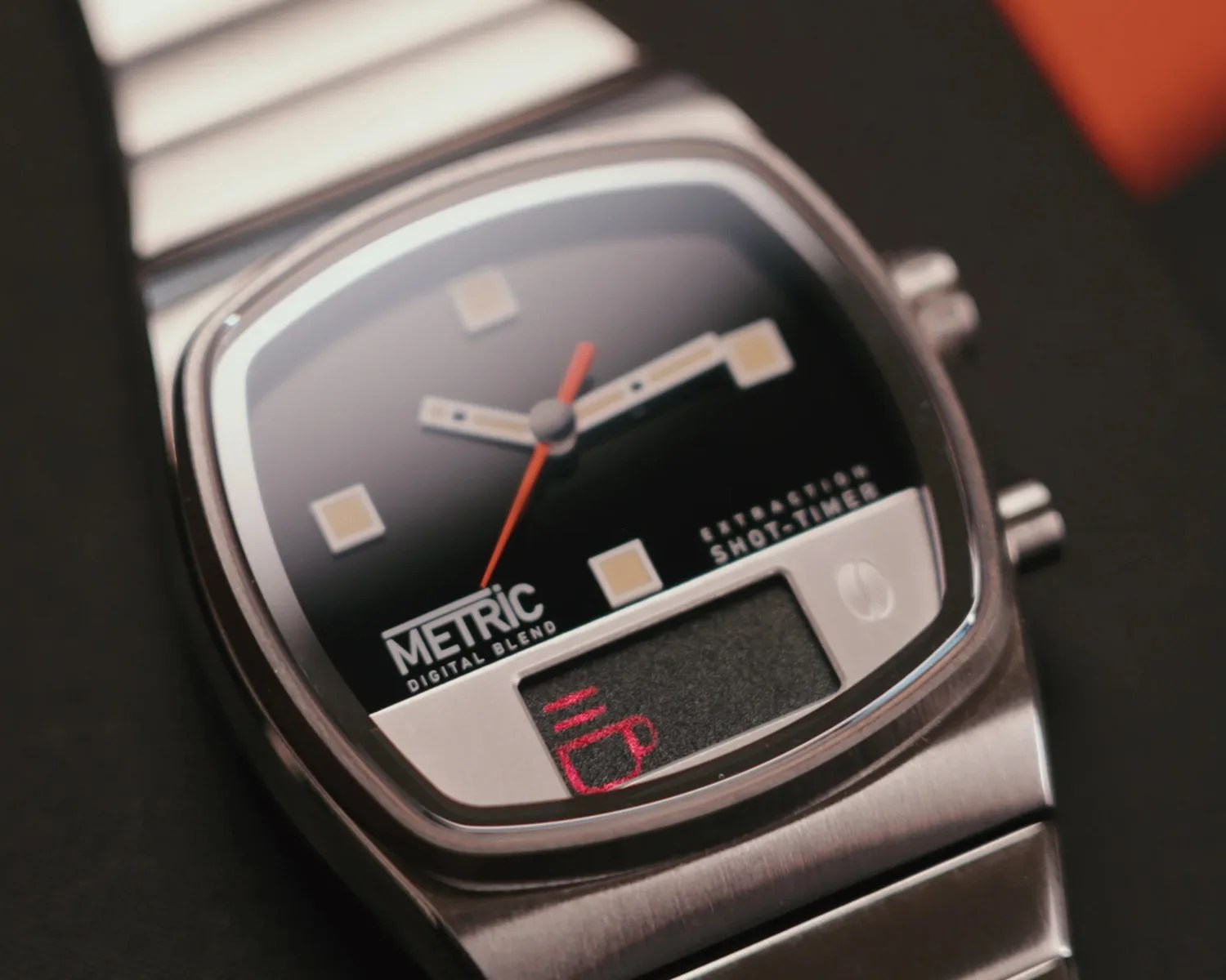Close-up of a silver wristwatch with a black dial, square hour markers, and a red second hand. The watch face displays the text "METRIC DIGITAL BLEND" and "EXTRACTION SHOT TIMER." There is a small digital screen at the bottom showing a red icon of a steaming coffee cup. The watch has a metal link band.