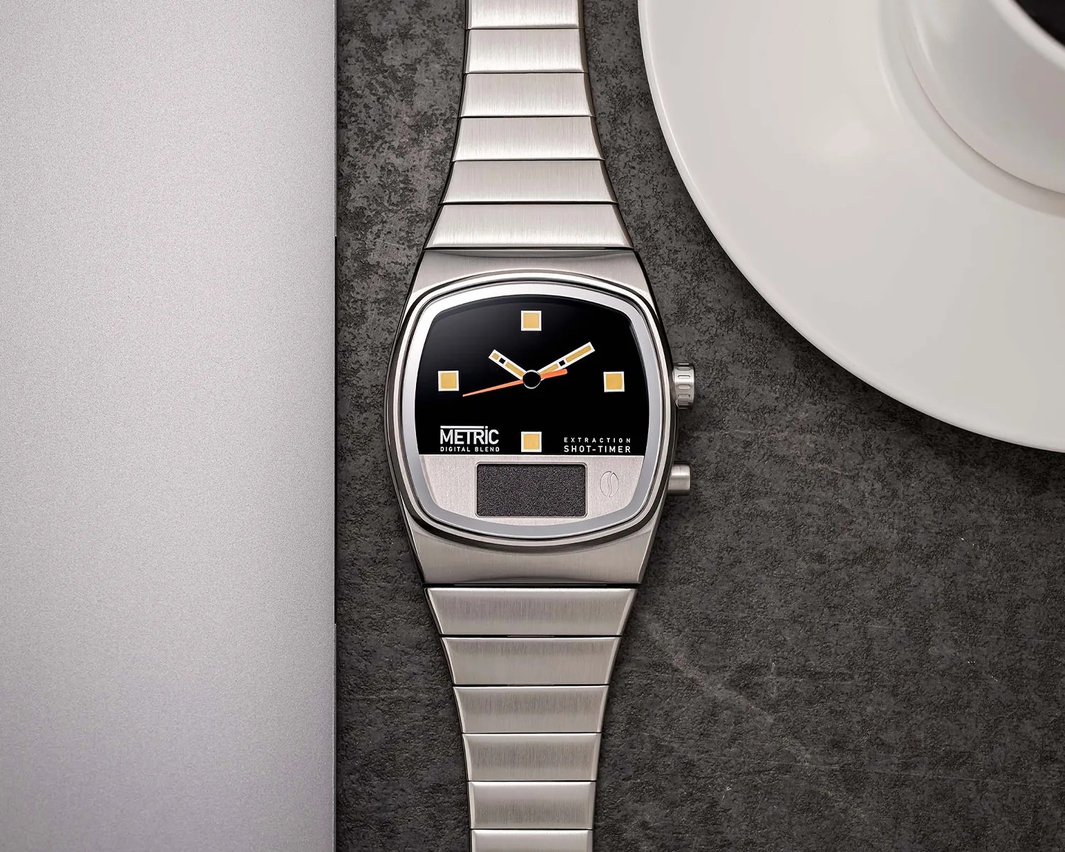 Silver metal wristwatch with a black and silver face, featuring square yellow hour markers and two yellow hands with black accents. The watch has a digital display area at the bottom and text reading "METRIC DIGITAL BLEND" and "EXTRACTION SHOT-TIMER." The watch is placed on a dark textured surface next to a silver laptop and a white coffee cup on a saucer.