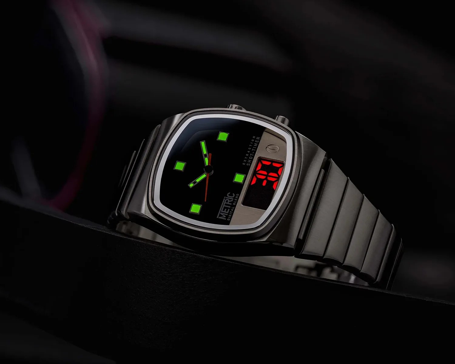 A stainless steel wristwatch with a black dial featuring green luminous square hour markers and hands. The watch has a digital red LED display on the right side and two push buttons on the top right of the case. The dial includes the text "METRIC DIGITAL BLEND" and "EXTRACTION SHOT TIMER." The watch band is a matching stainless steel link bracelet.