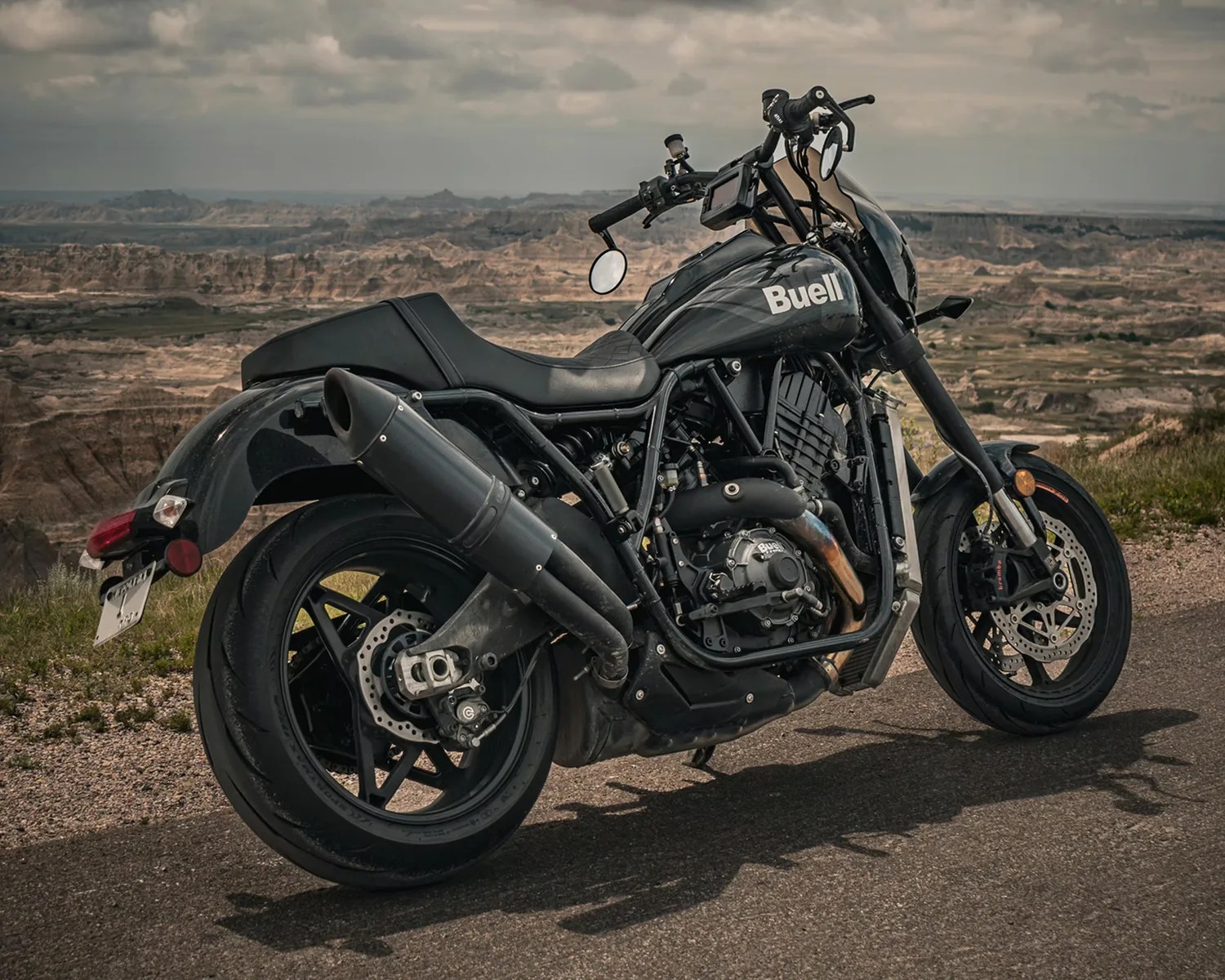 Black Buell motorcycle parked on a paved road with a rugged, rocky landscape in the background under a cloudy sky. The motorcycle features a large exhaust pipe, disc brakes, and a comfortable seat.