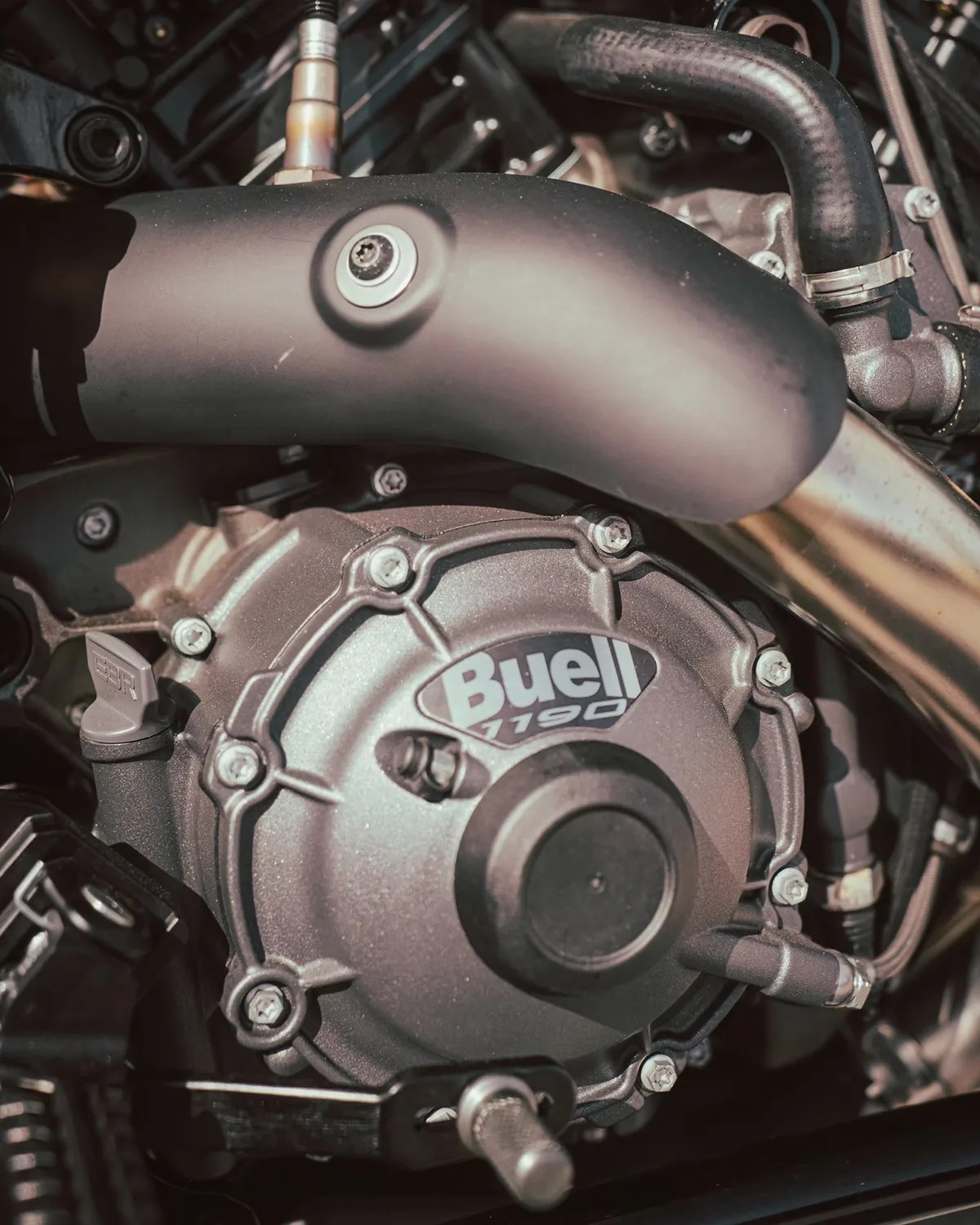 Close-up of a Buell 1190 motorcycle engine, showing the metallic engine casing with the "Buell 1190" logo prominently displayed. The image also features various bolts, a black exhaust pipe, and connected hoses.