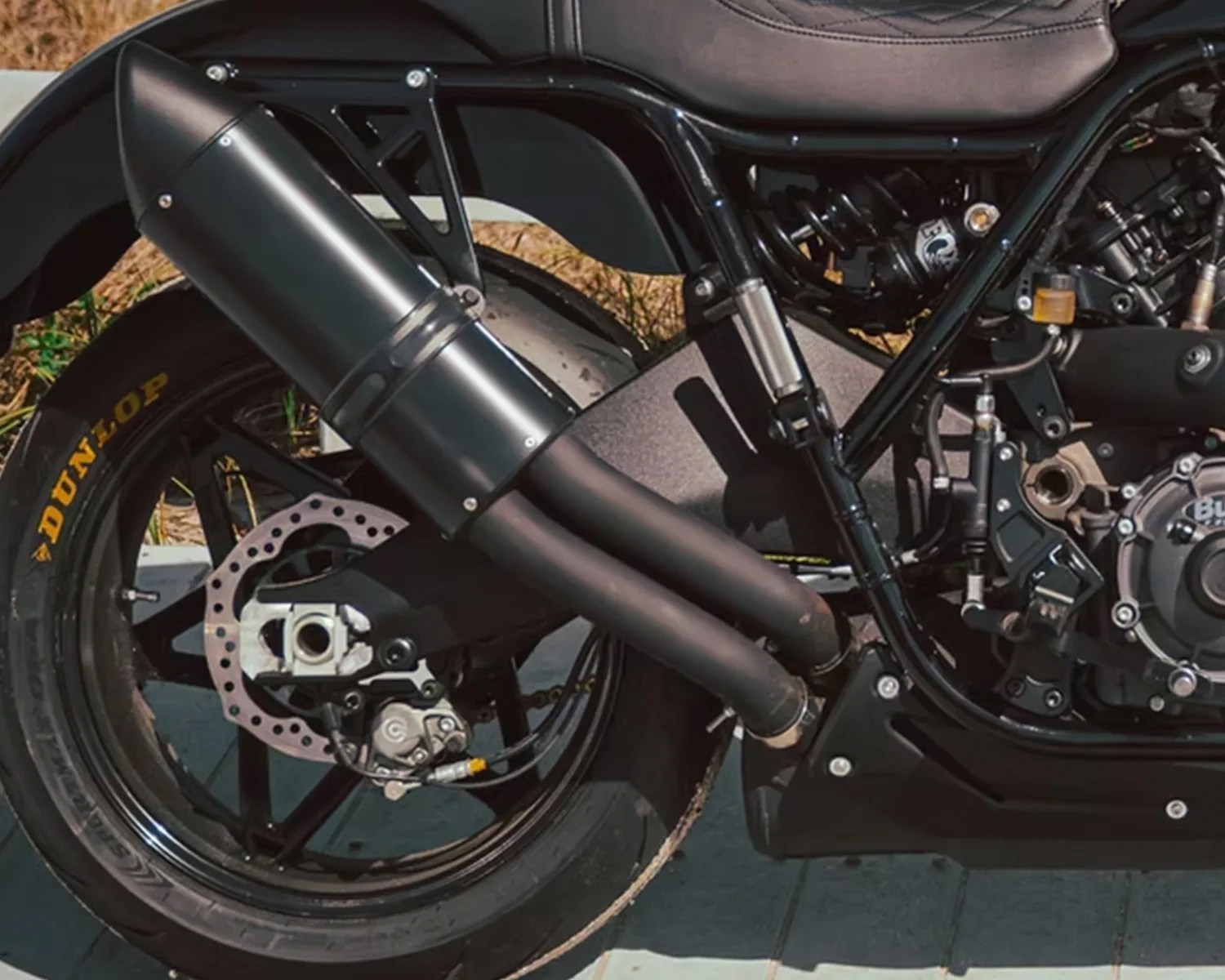 Close-up of the rear section of a motorcycle showing the black exhaust pipes, rear wheel with Dunlop tire, disc brake, and part of the engine and frame. The seat and suspension components are also partially visible.