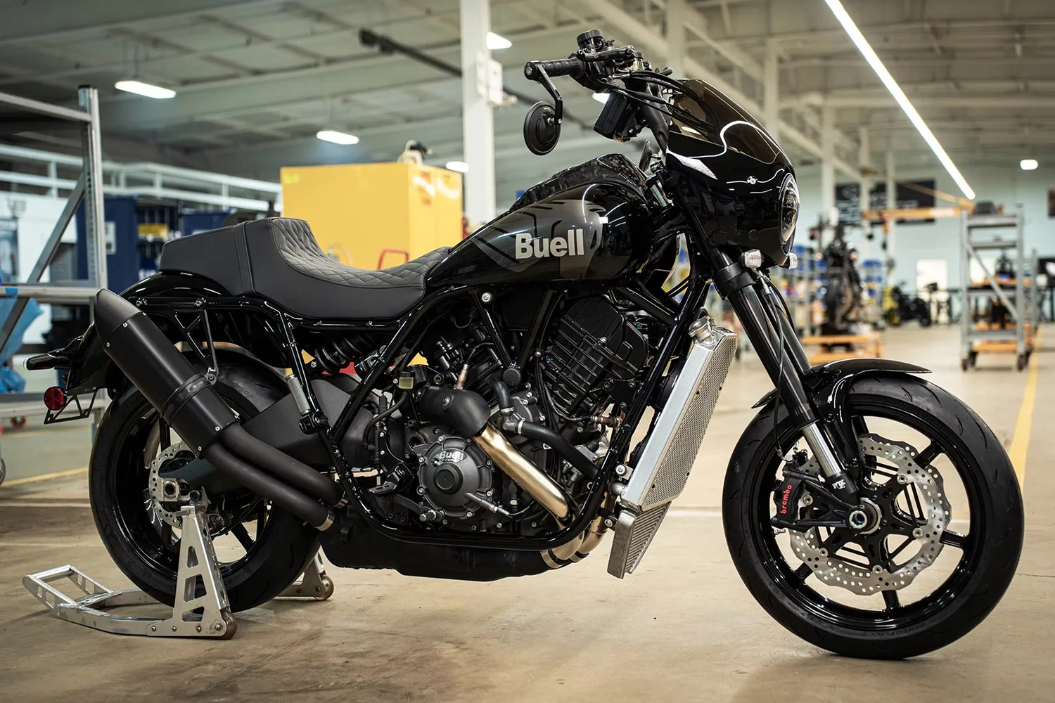 Black Buell motorcycle with a quilted seat, dual exhaust pipes, and a front fairing, parked indoors on a stand in a workshop setting.