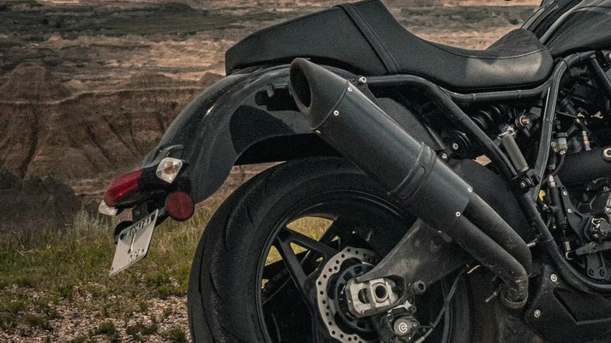 Rear section of a black motorcycle showing the rear tire, exhaust pipes, brake disc, and part of the seat, with a natural rocky landscape in the background.