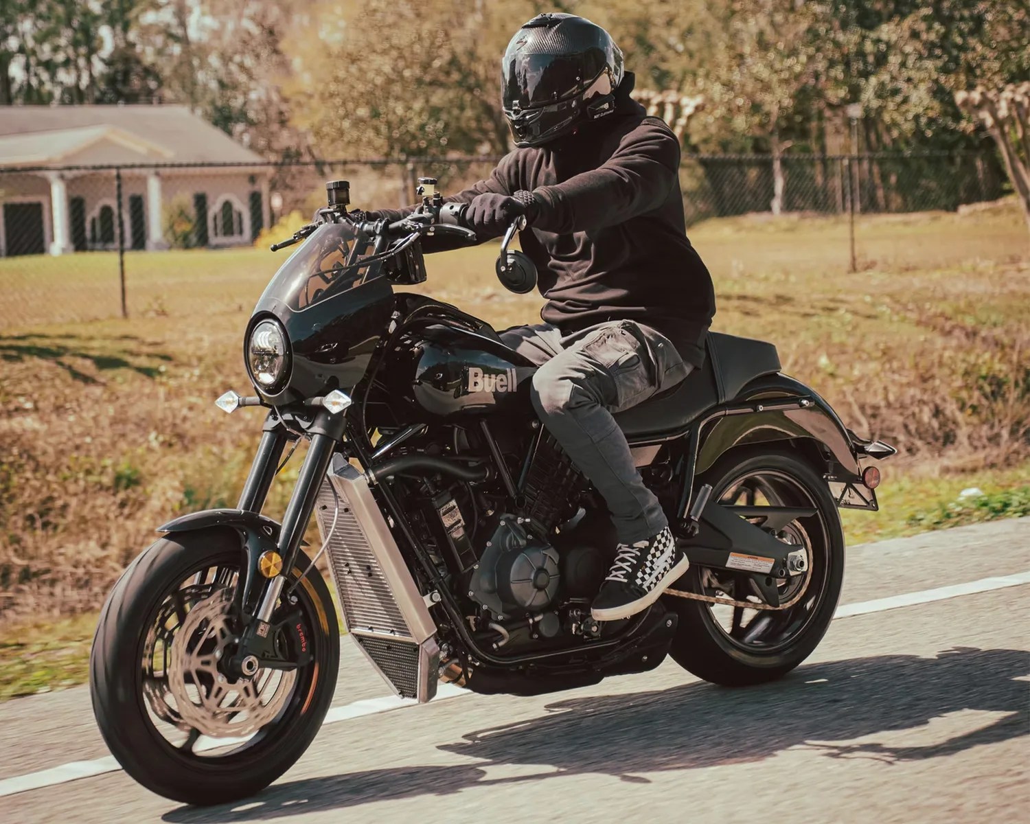 A person wearing a black helmet, black hoodie, gray cargo pants, and black-and-white checkered sneakers is riding a black Buell motorcycle on a paved road with a grassy area and trees in the background.