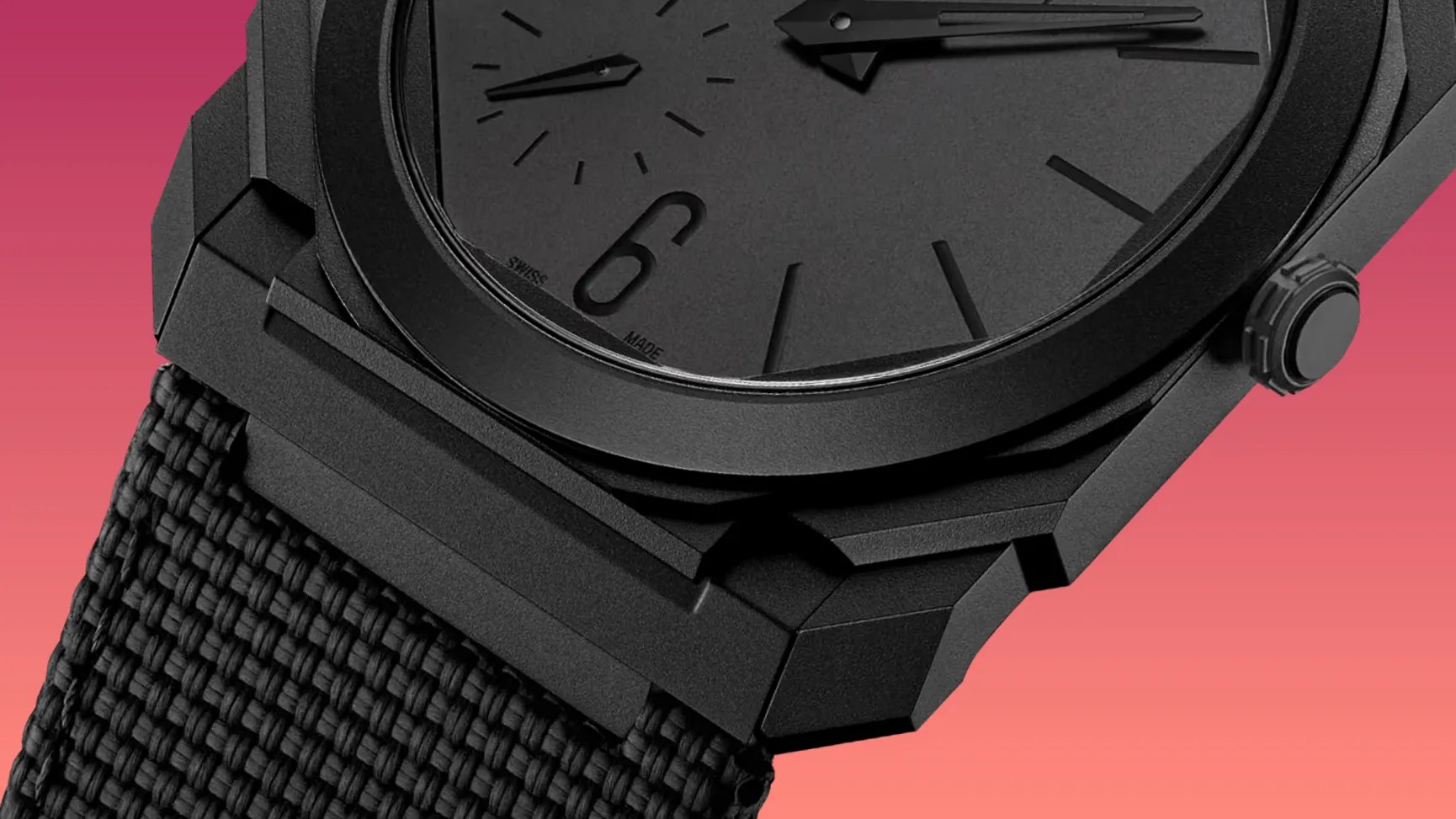 Close-up of a black wristwatch with a matte finish, featuring a textured black fabric strap. The watch face is black with minimalistic hour markers and hands, and the number 6 is prominently displayed. The crown is also black and textured for grip. The background is a gradient of pink to orange.