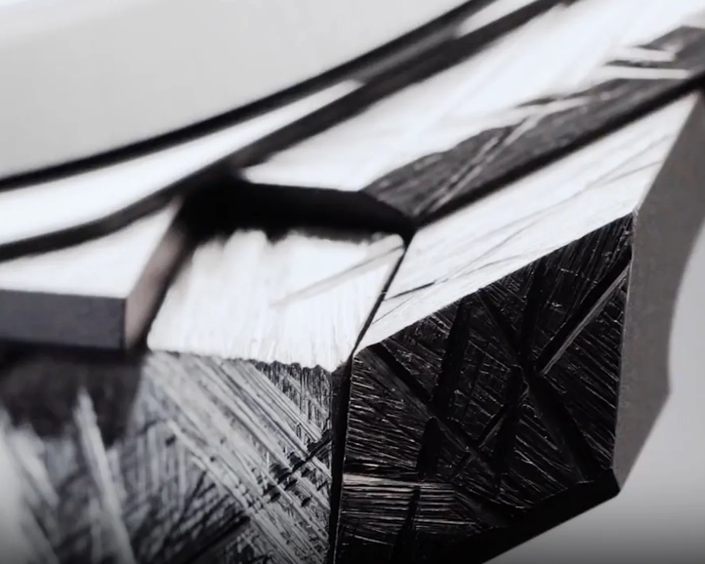 Close-up of a metallic, faceted surface with sharp edges and a reflective finish, showing fine linear textures and scratches on the darker areas. The image highlights the contrast between polished and rough sections.